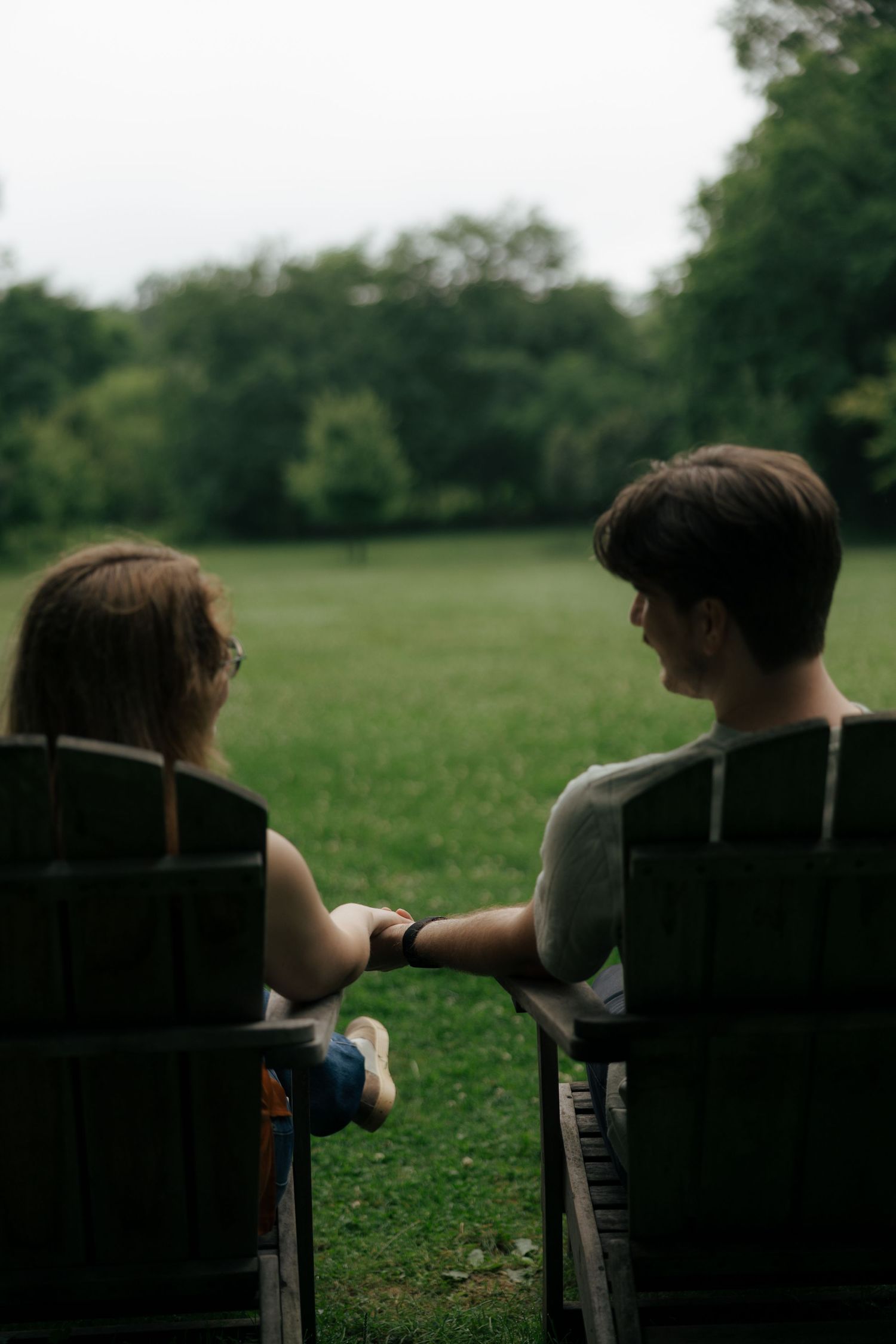 Couple sits in wooden Adirondack chairs holding hands while overlooking a serene green park landscape.