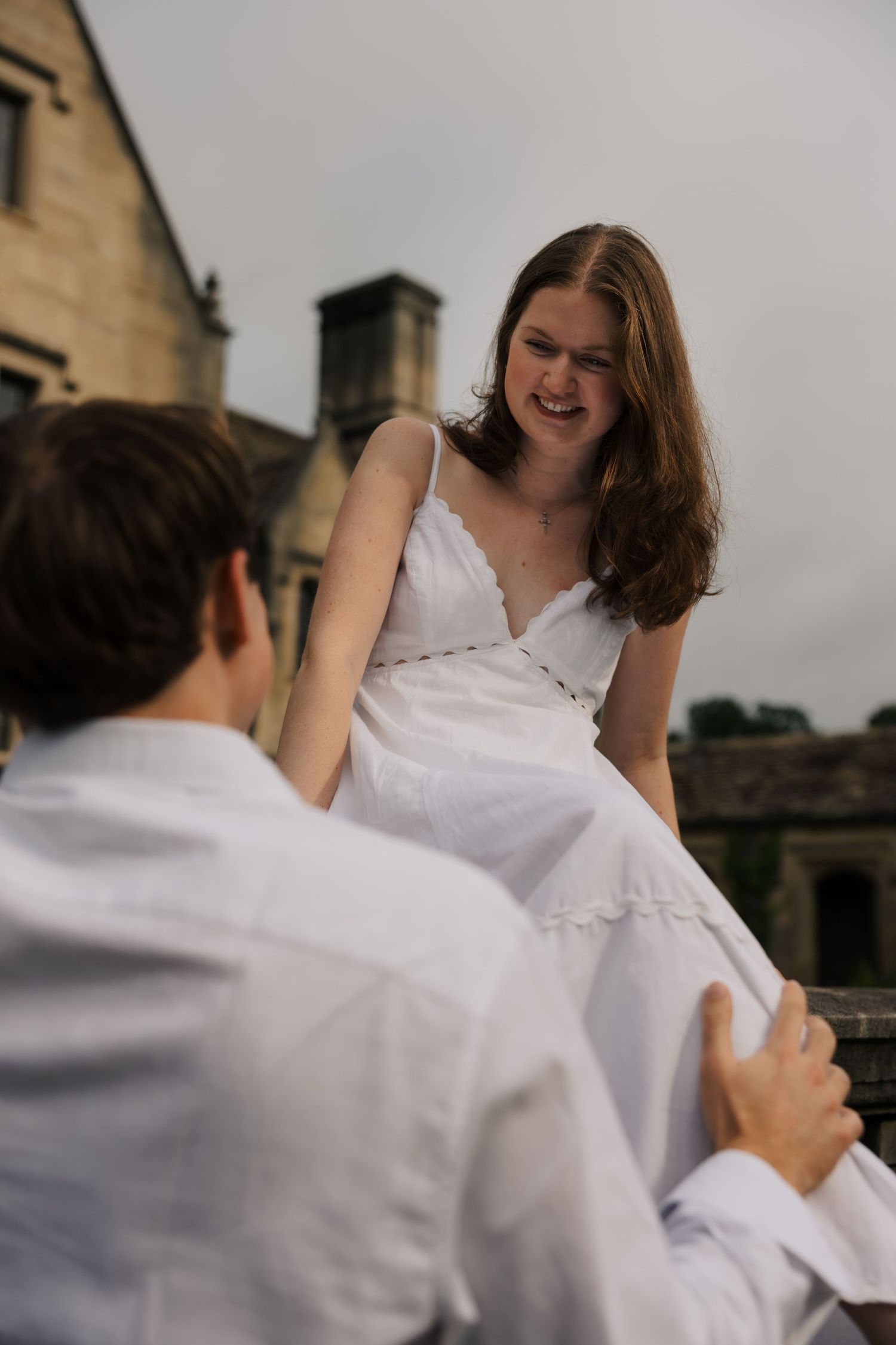 Smiling woman in white dress sits on stone ledge reaching toward her partner outdoors.
