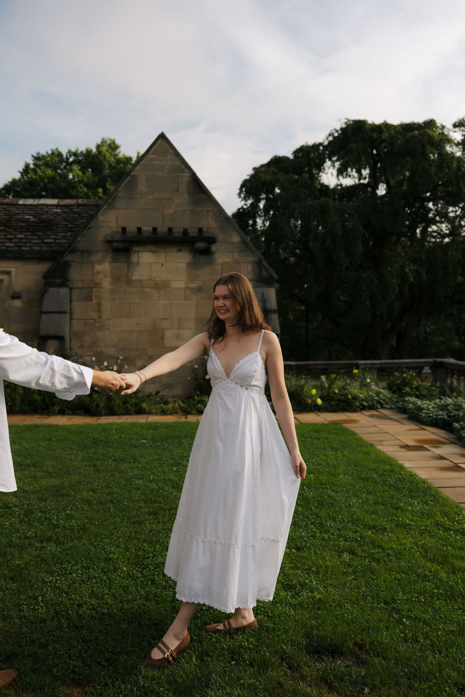 Woman in white sundress holds hands with partner on green lawn before stone manor house.