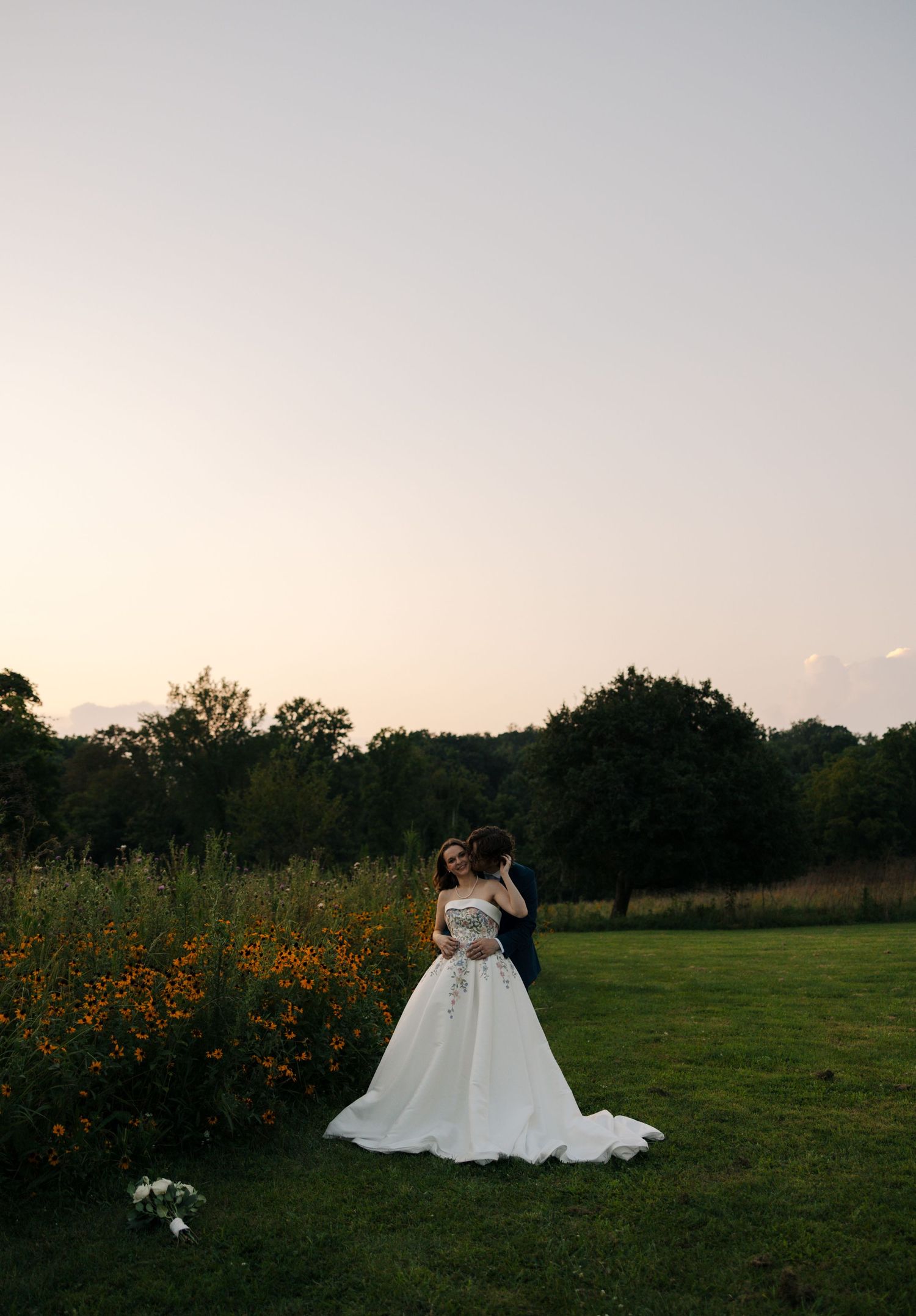 Bride and groom embrace at sunset in a lush green field with orange wildflowers and trees behind them.