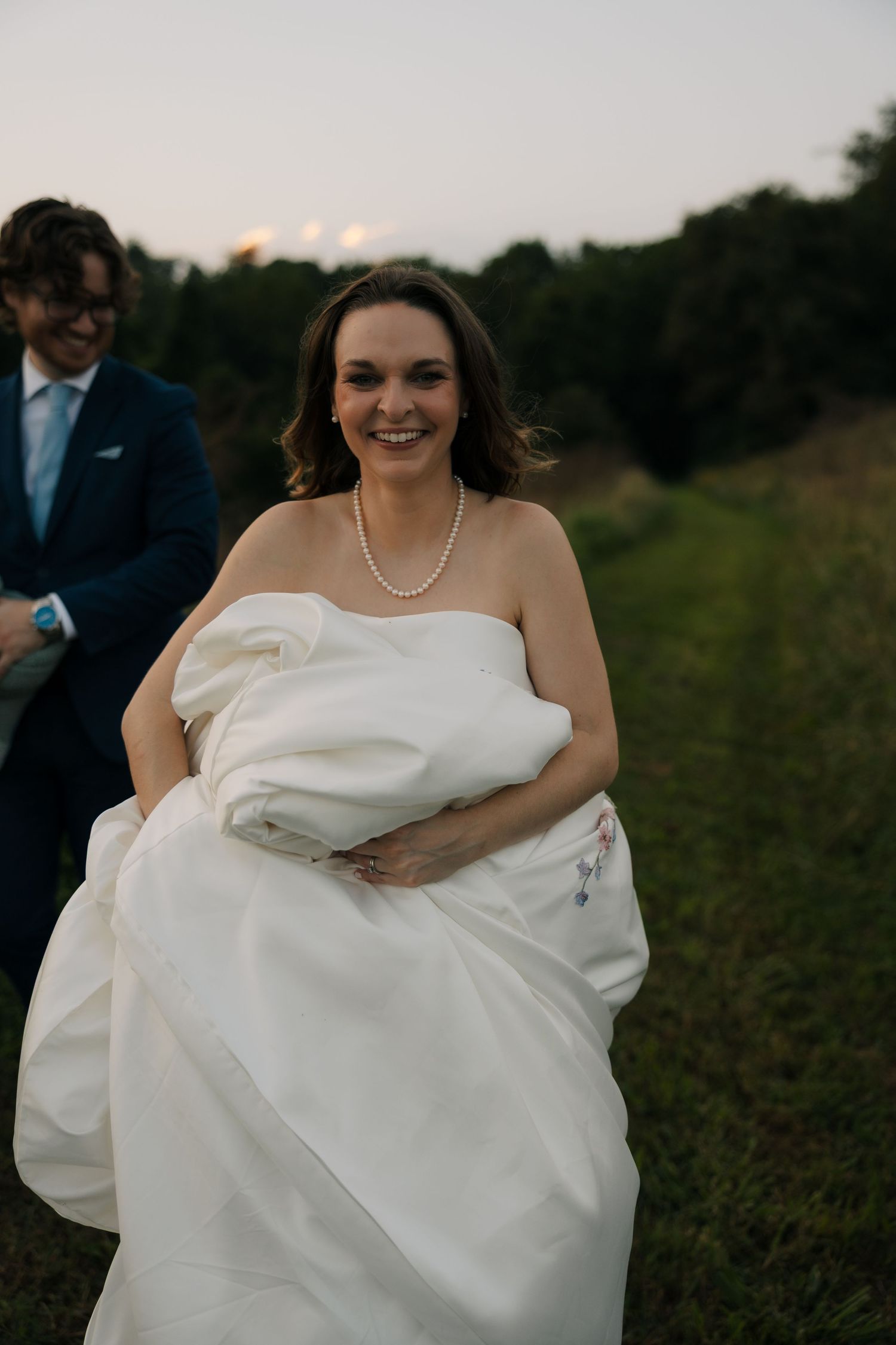 Smiling bride in strapless white gown holds her dress while walking through a grassy outdoor setting.