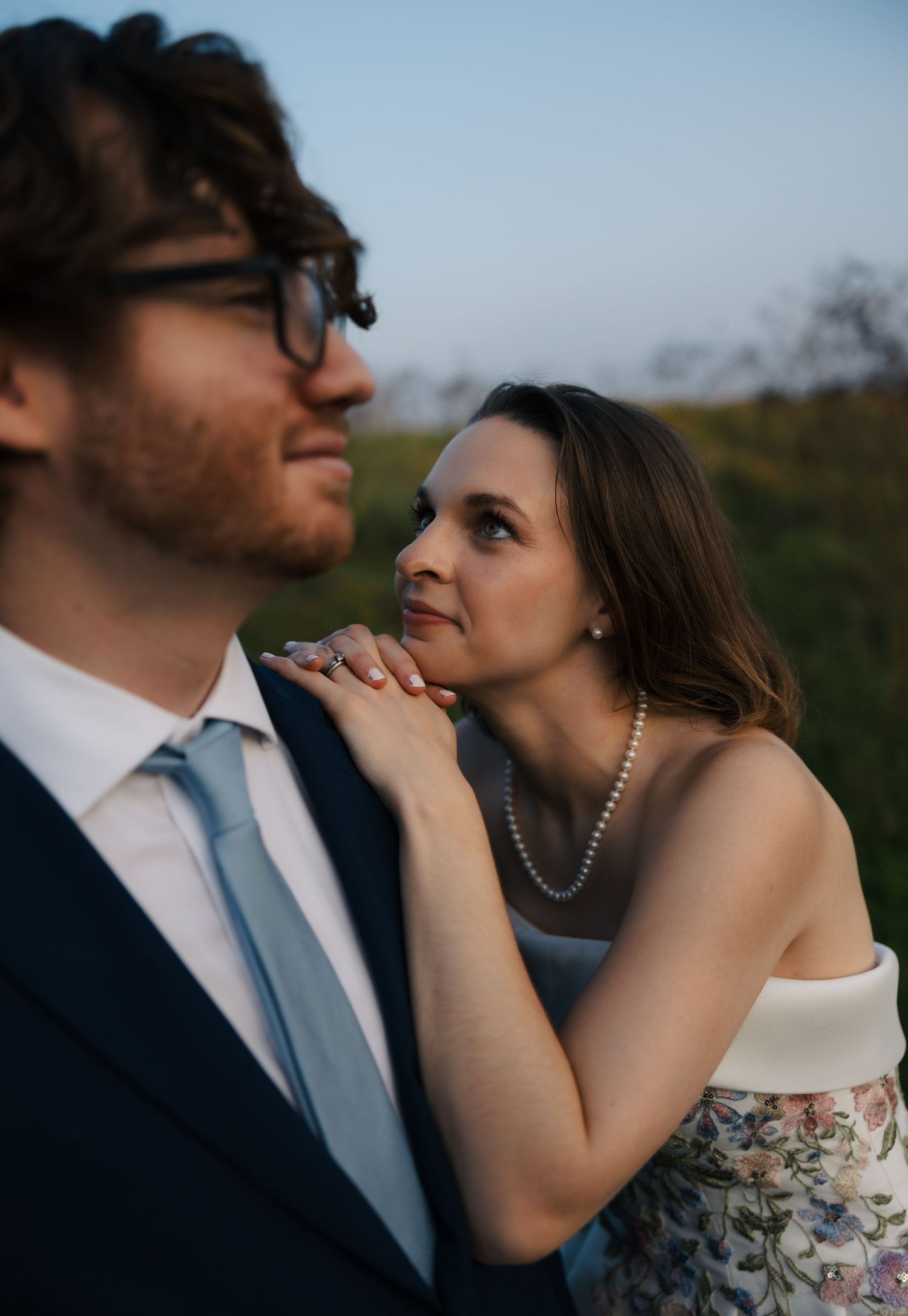 Bride in floral strapless gown with pearl necklace looks adoringly at groom in navy suit at golden hour.