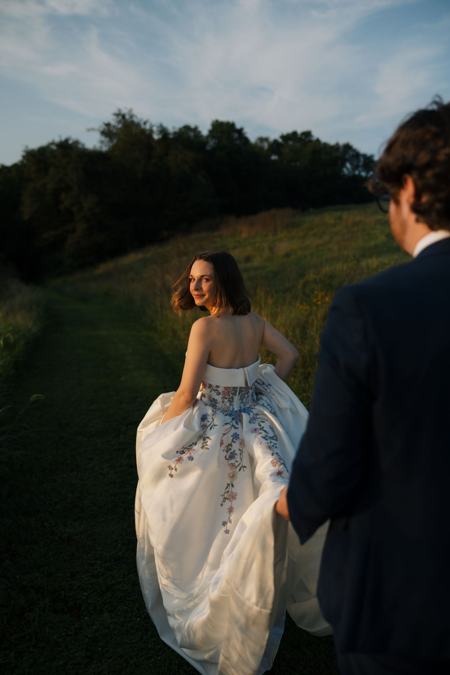 Bride in floral embroidered white gown glances back at groom while walking through lush green meadow at golden hour.