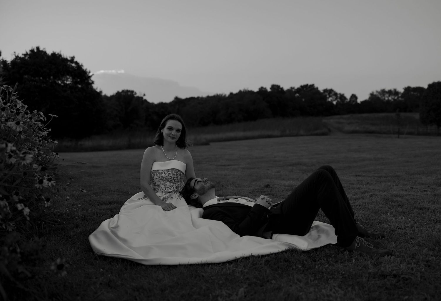 Black and white photo of bride and groom relaxing on lawn at twilight, groom lying across brides lap.