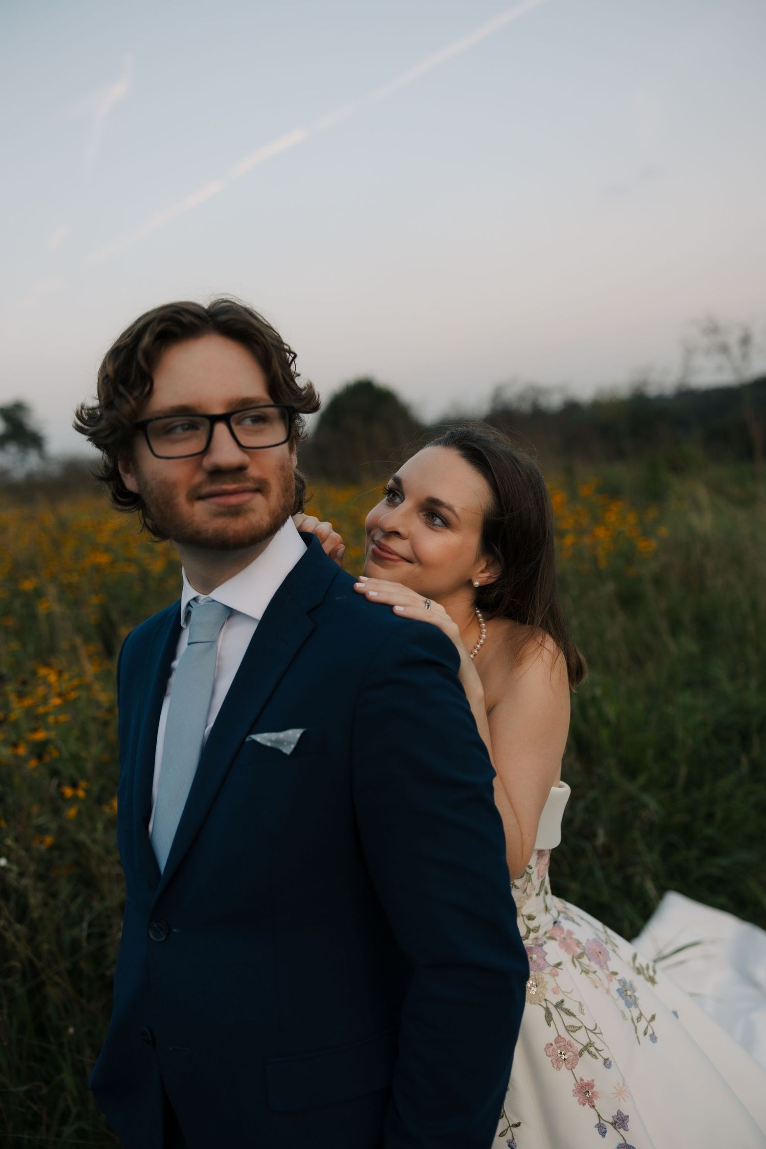 Bride in floral wedding dress embraces groom in navy suit from behind in a golden wildflower meadow.