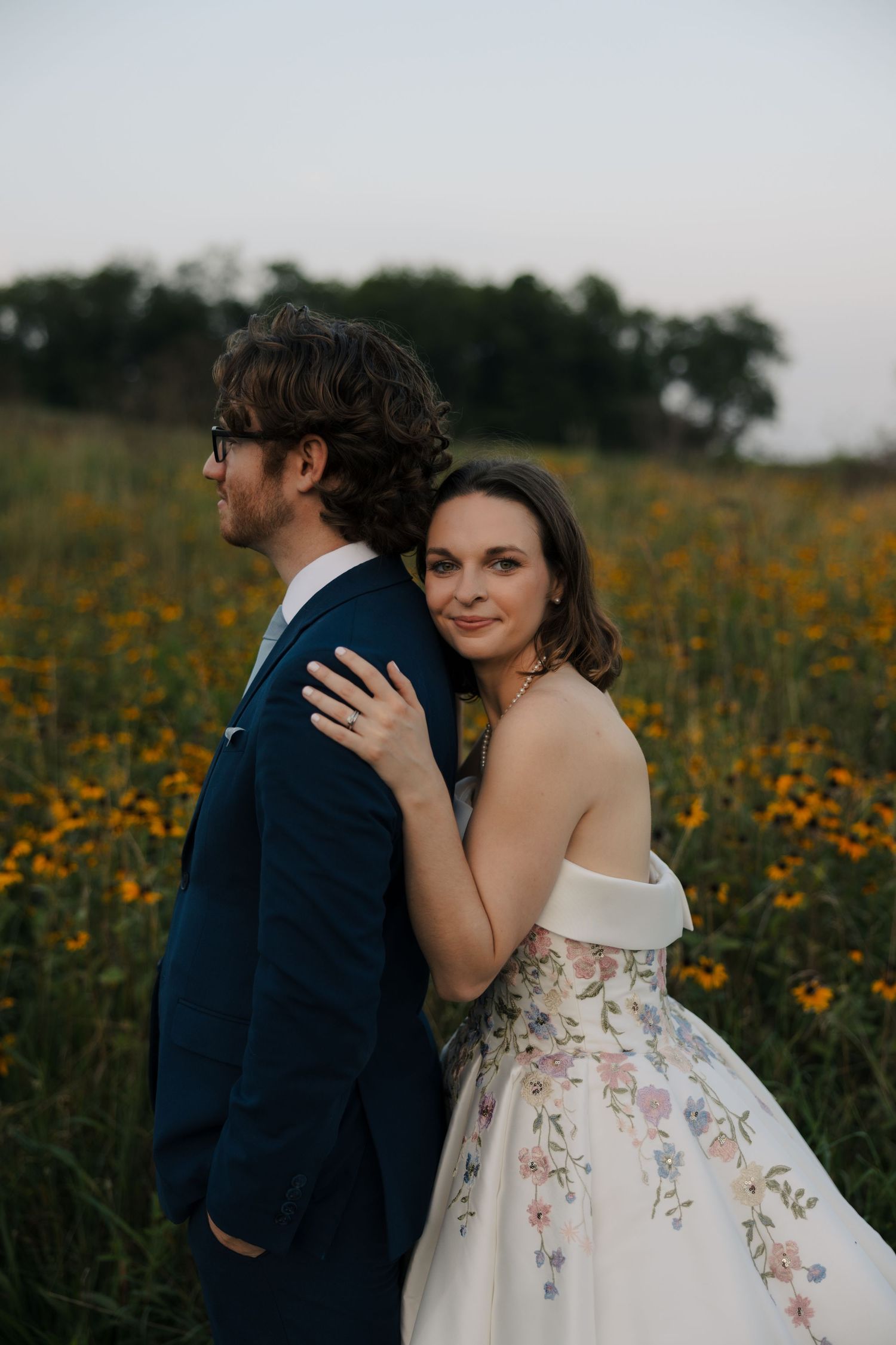Bride in off-shoulder floral gown leans on groom's back in wildflower field at dusk wedding portrait.
