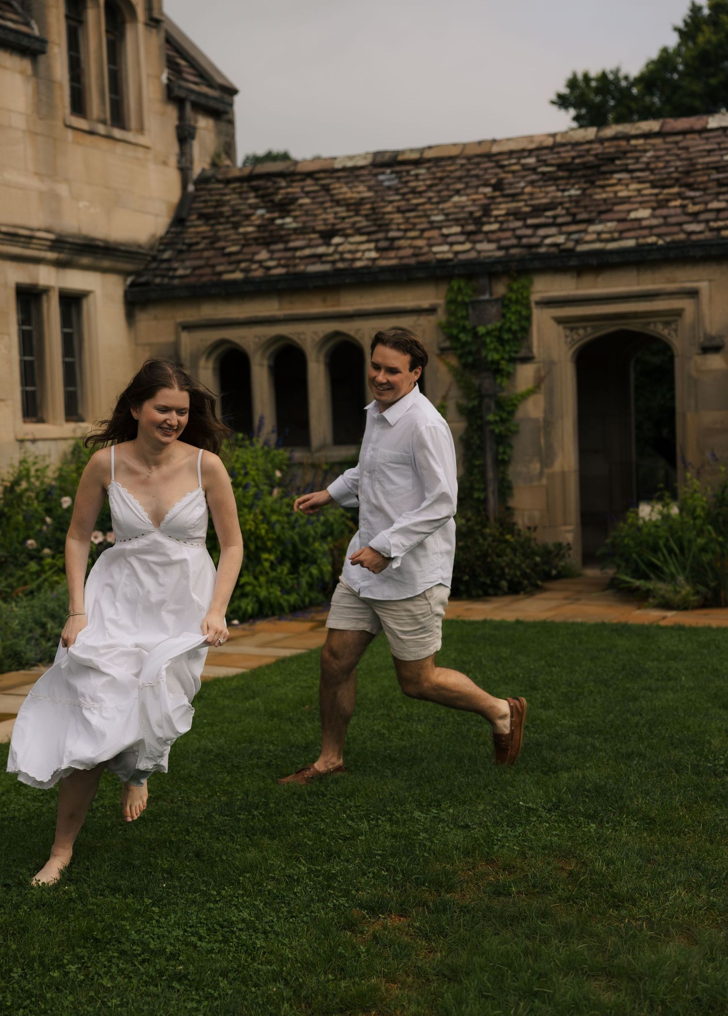 Joyful couple running barefoot on green lawn outside historic stone manor house on cloudy day.