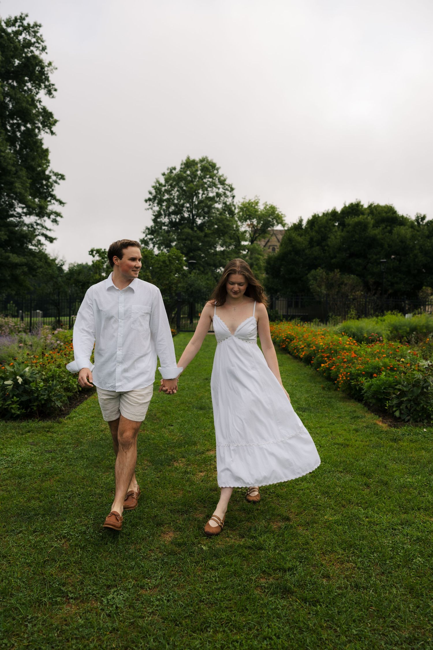 Couple in matching white outfits holding hands while walking through a lush garden path lined with colorful flowers.