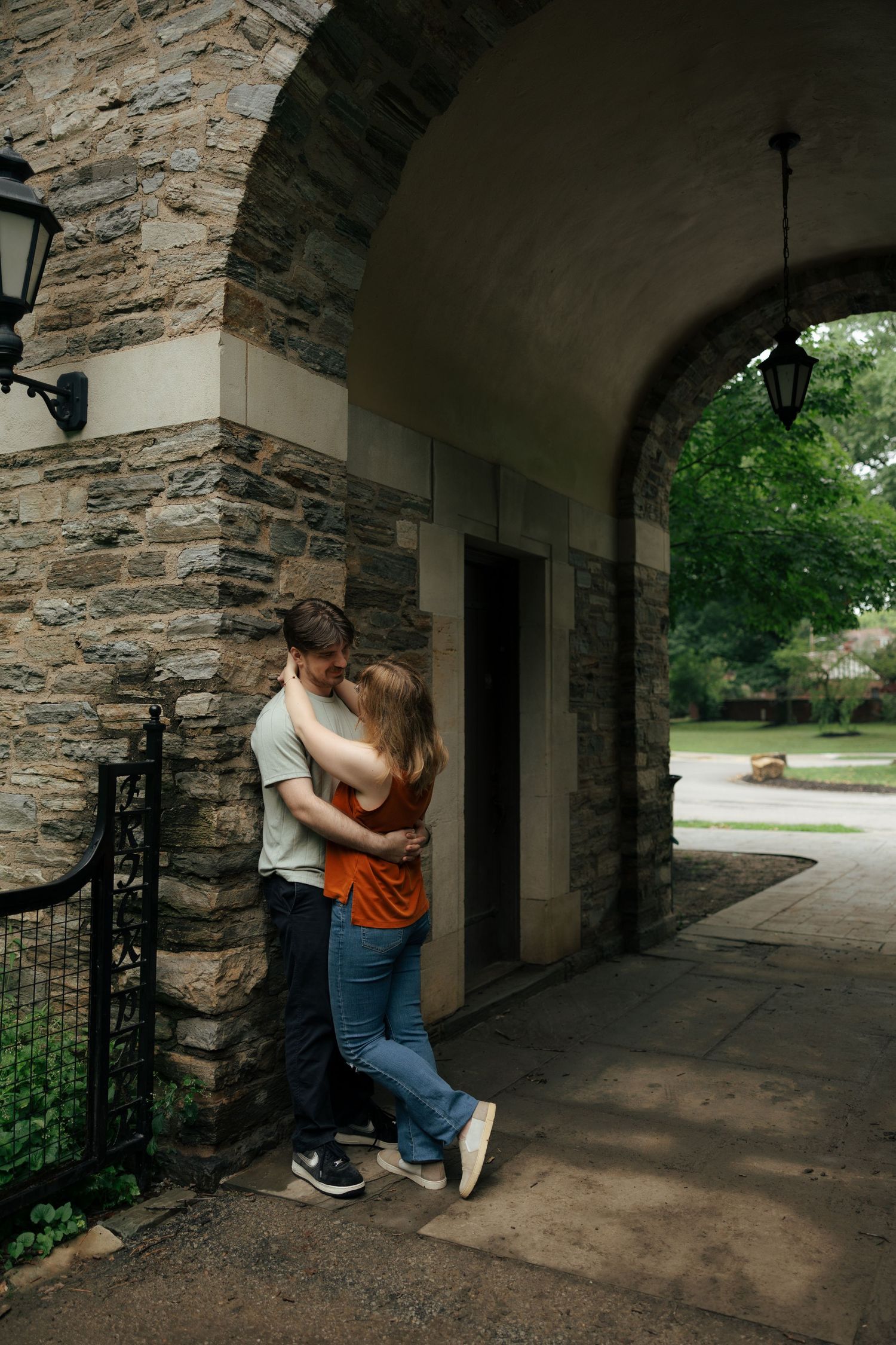 Couple kissing against a stone wall under an arched entryway in a scenic outdoor park setting.