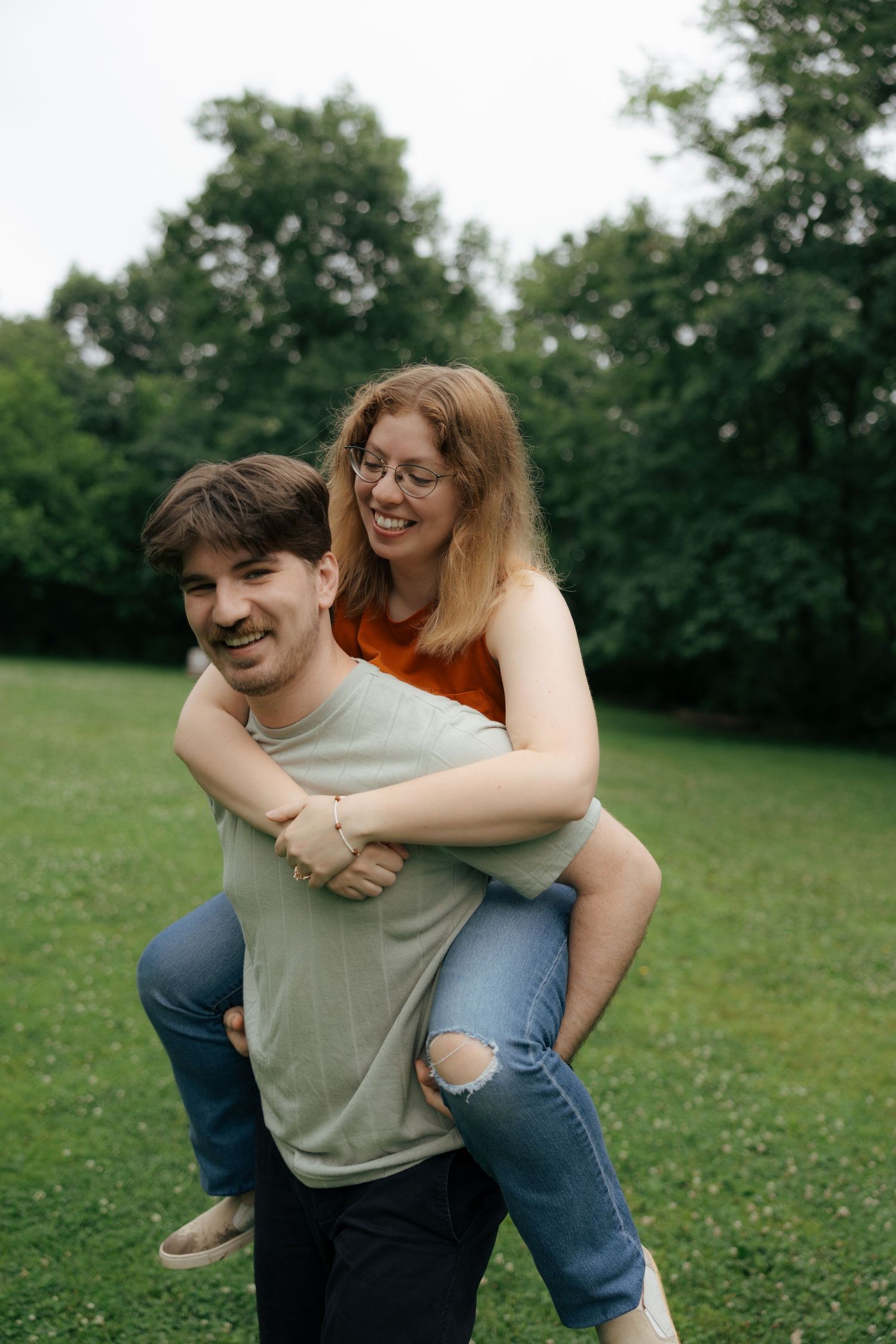 A man gives a smiling woman a piggyback ride in a lush green park on a cloudy day.