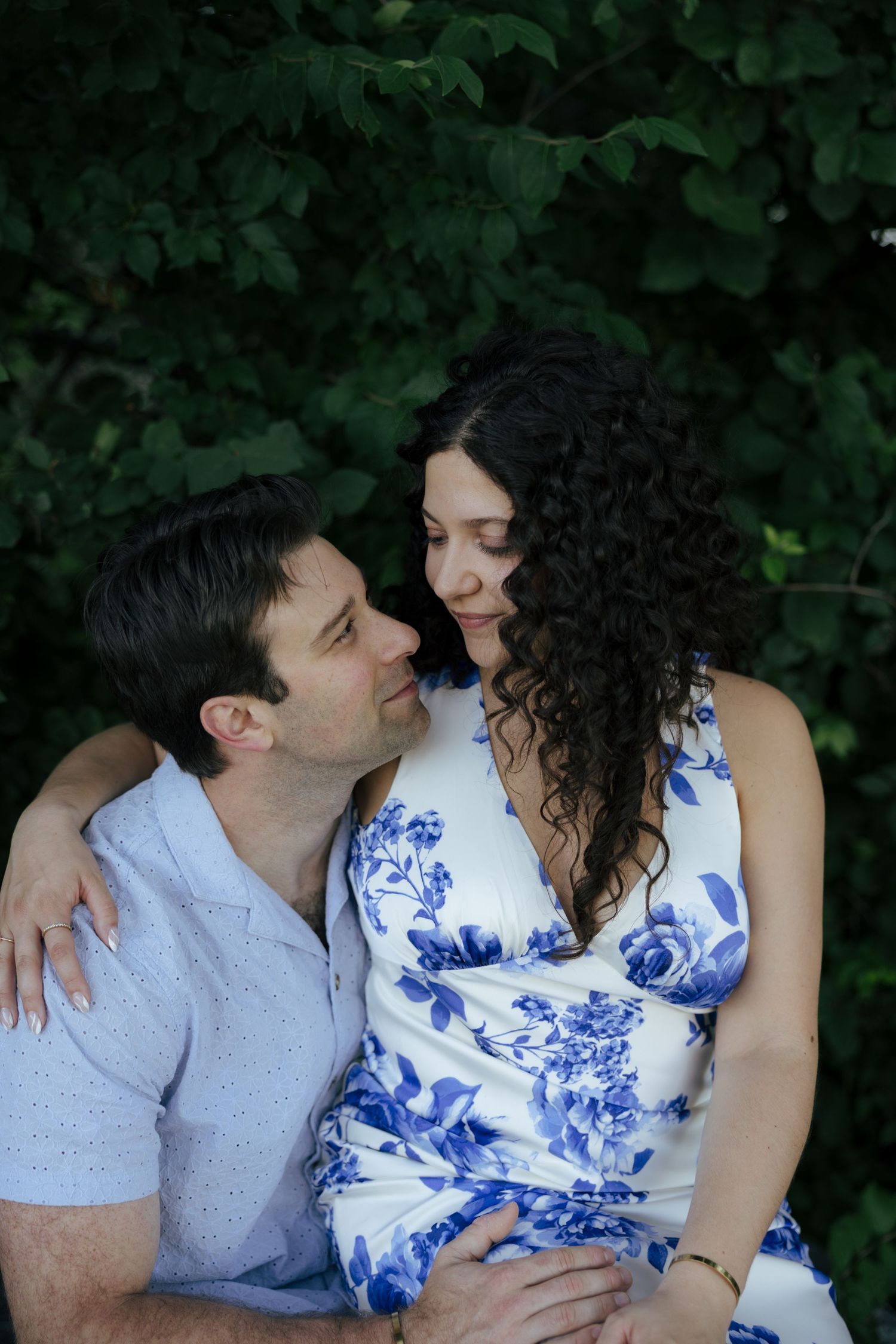 Couple sharing a romantic moment outdoors, woman in blue floral dress embraced by man in light blue shirt against green foliage.