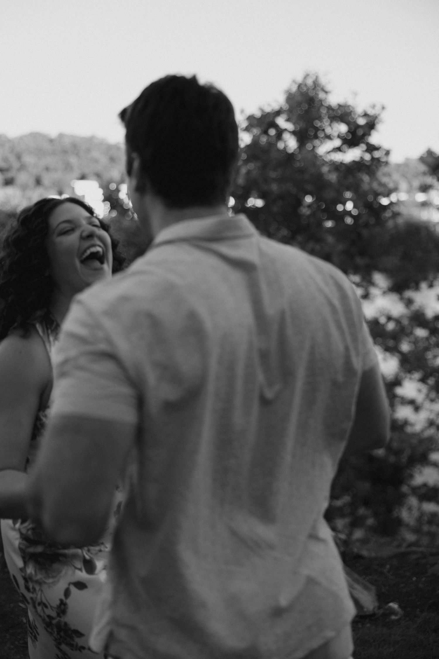 Black and white photo of a laughing woman holding hands with her partner outdoors among blurred trees.