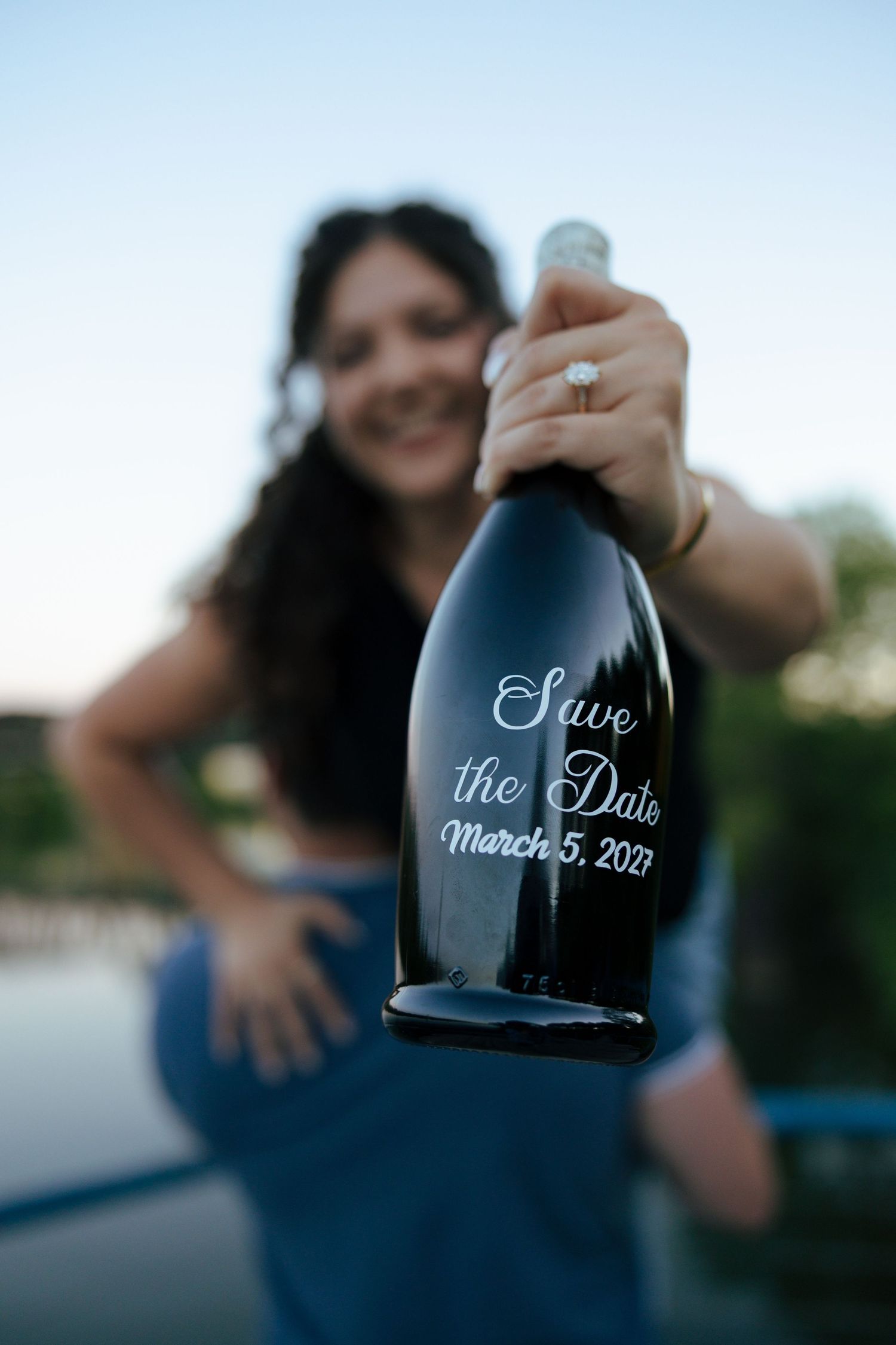 Woman holding engraved Save the Date bottle for March 5, 2027 engagement announcement outdoors.