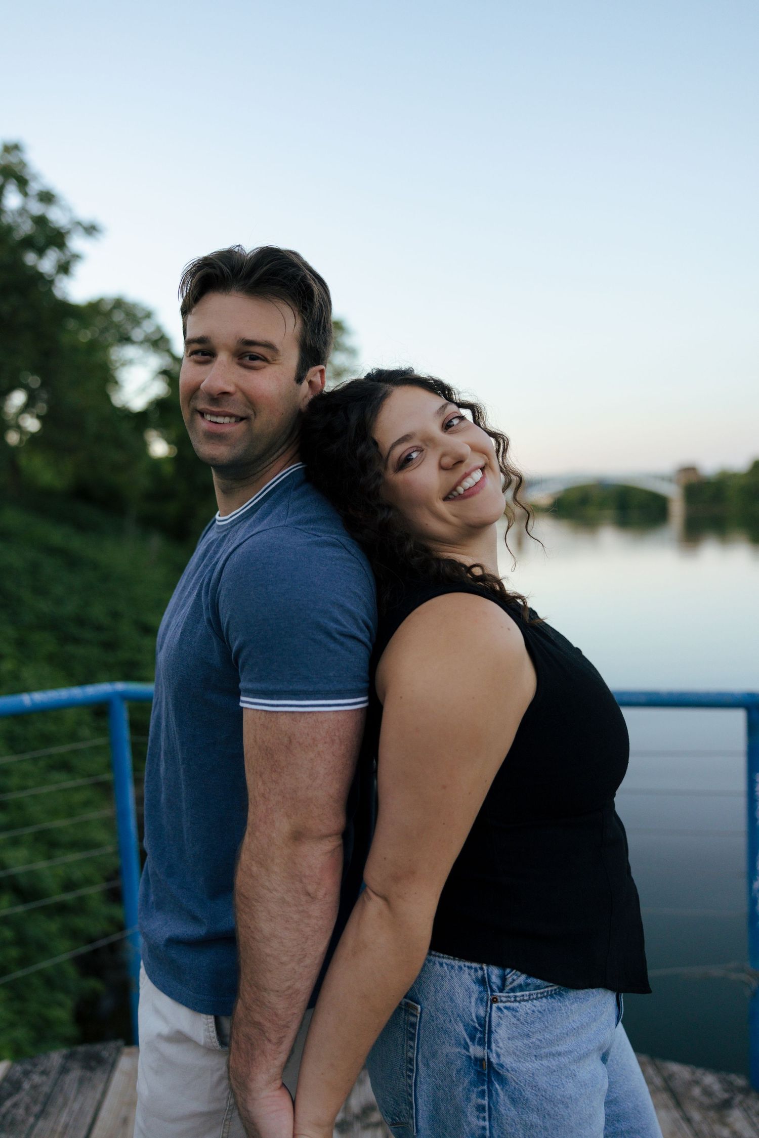 A smiling couple stands back-to-back on a bridge overlooking a serene river surrounded by green trees.