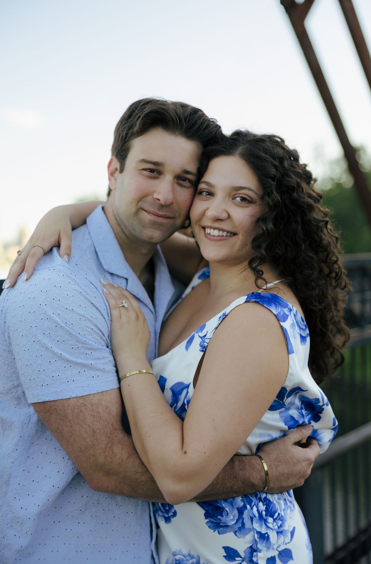 Smiling couple embracing on bridge, man in light blue shirt and woman in blue floral dress.