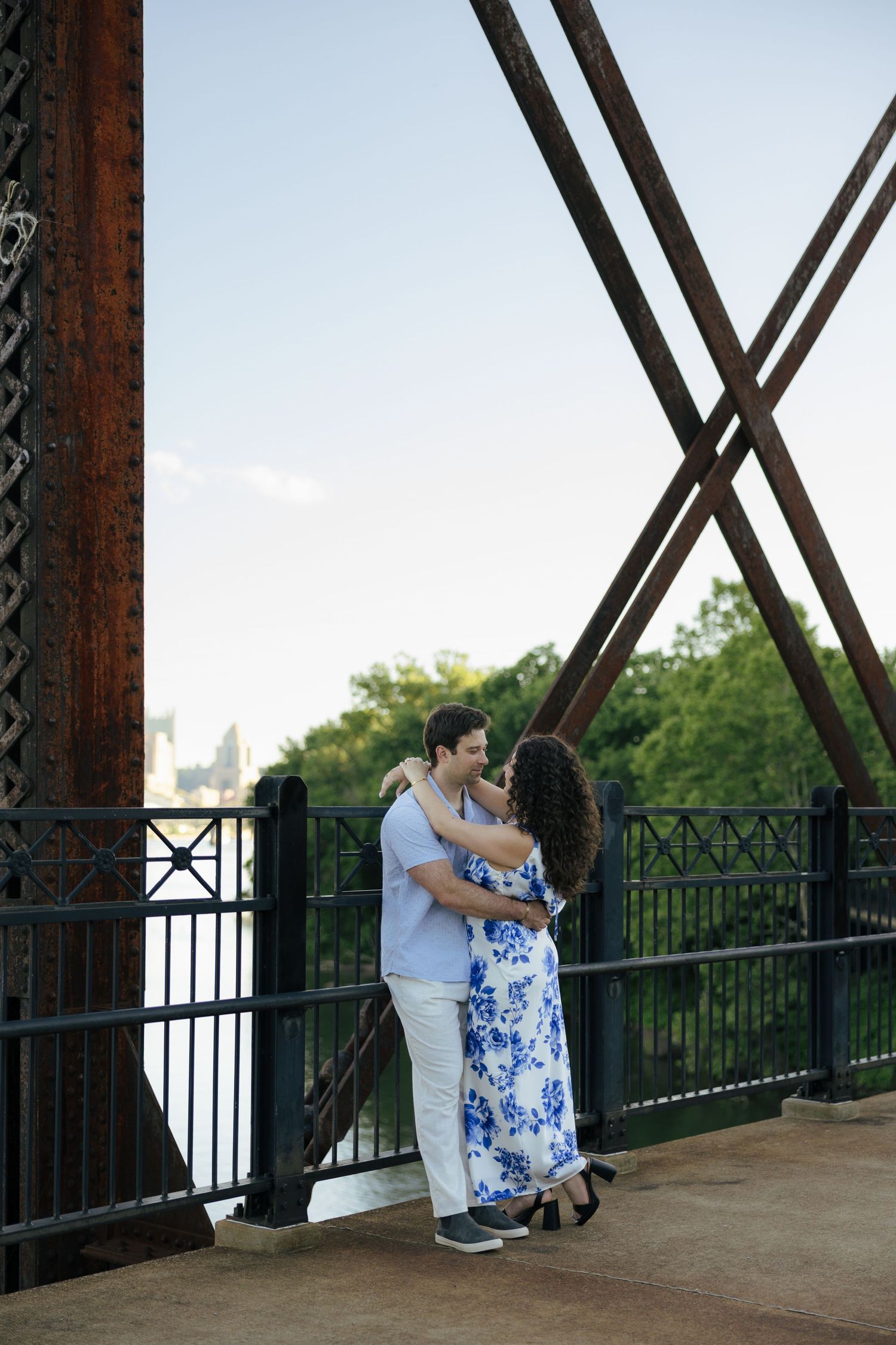 A couple embraces and smiles at each other on a historic iron bridge with a river view at sunset.