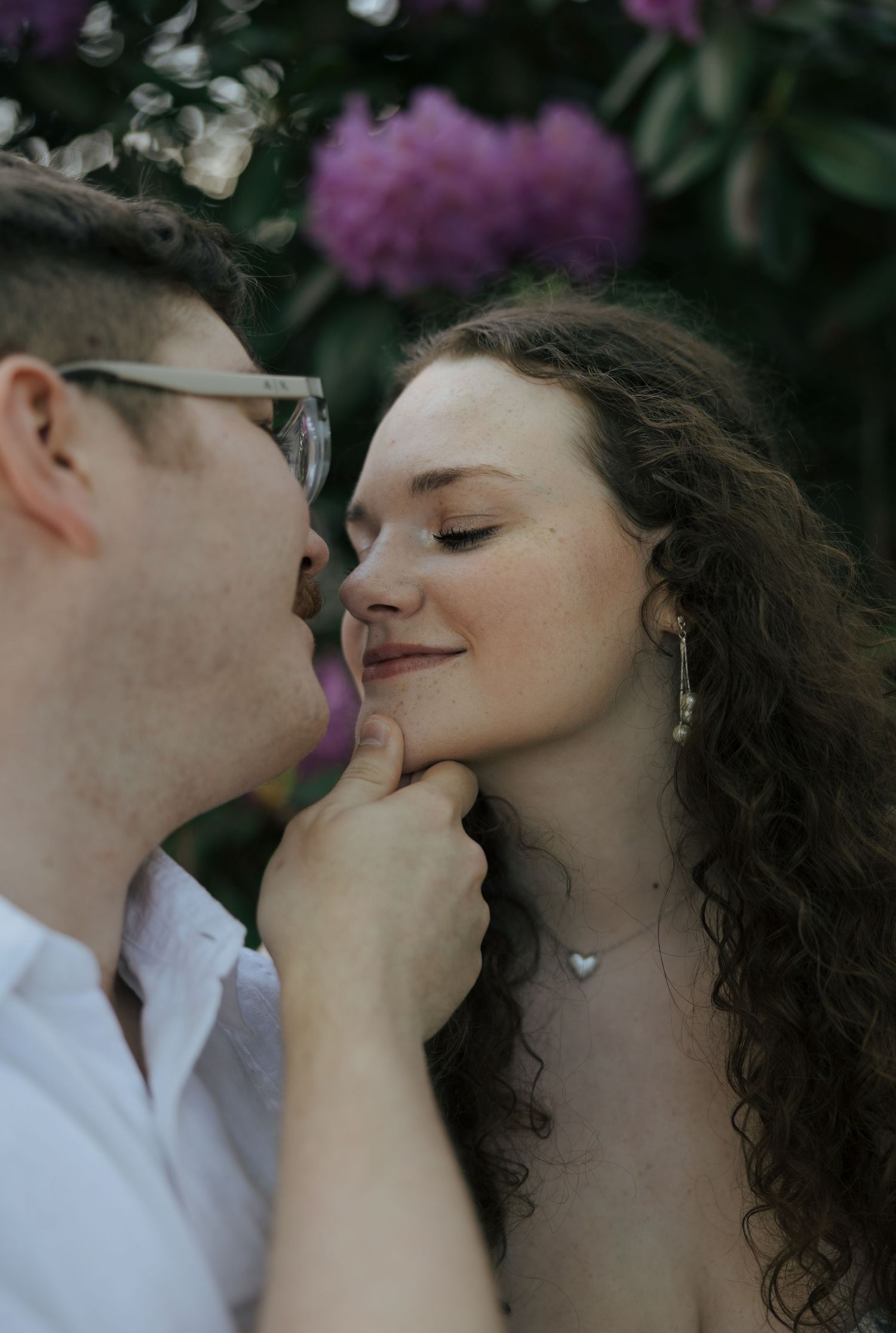 Couple sharing an intimate moment near purple flowers, woman smiling with eyes closed.
