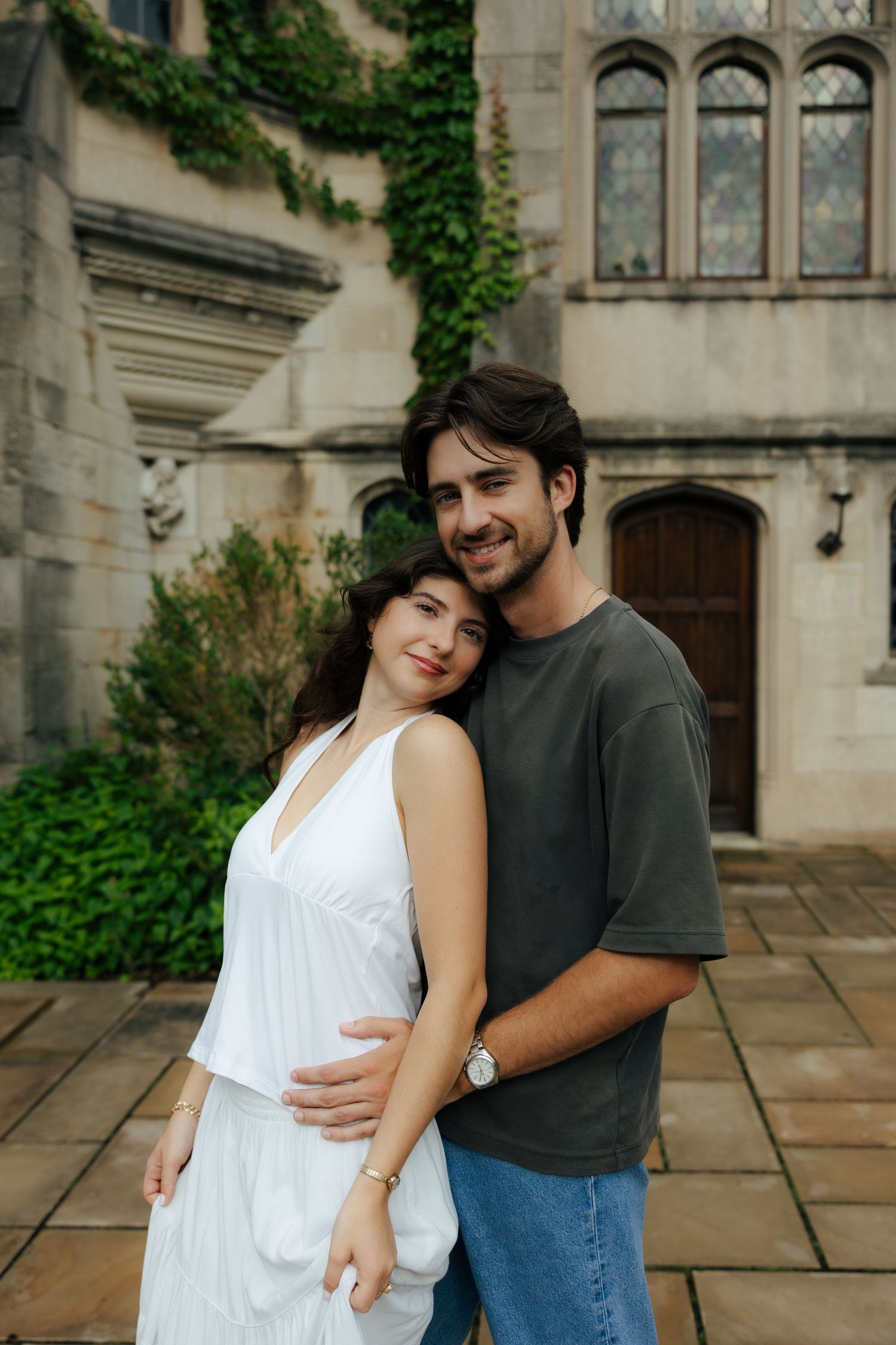 A happy couple embraces in front of an ivy-covered stone building, woman in white dress.