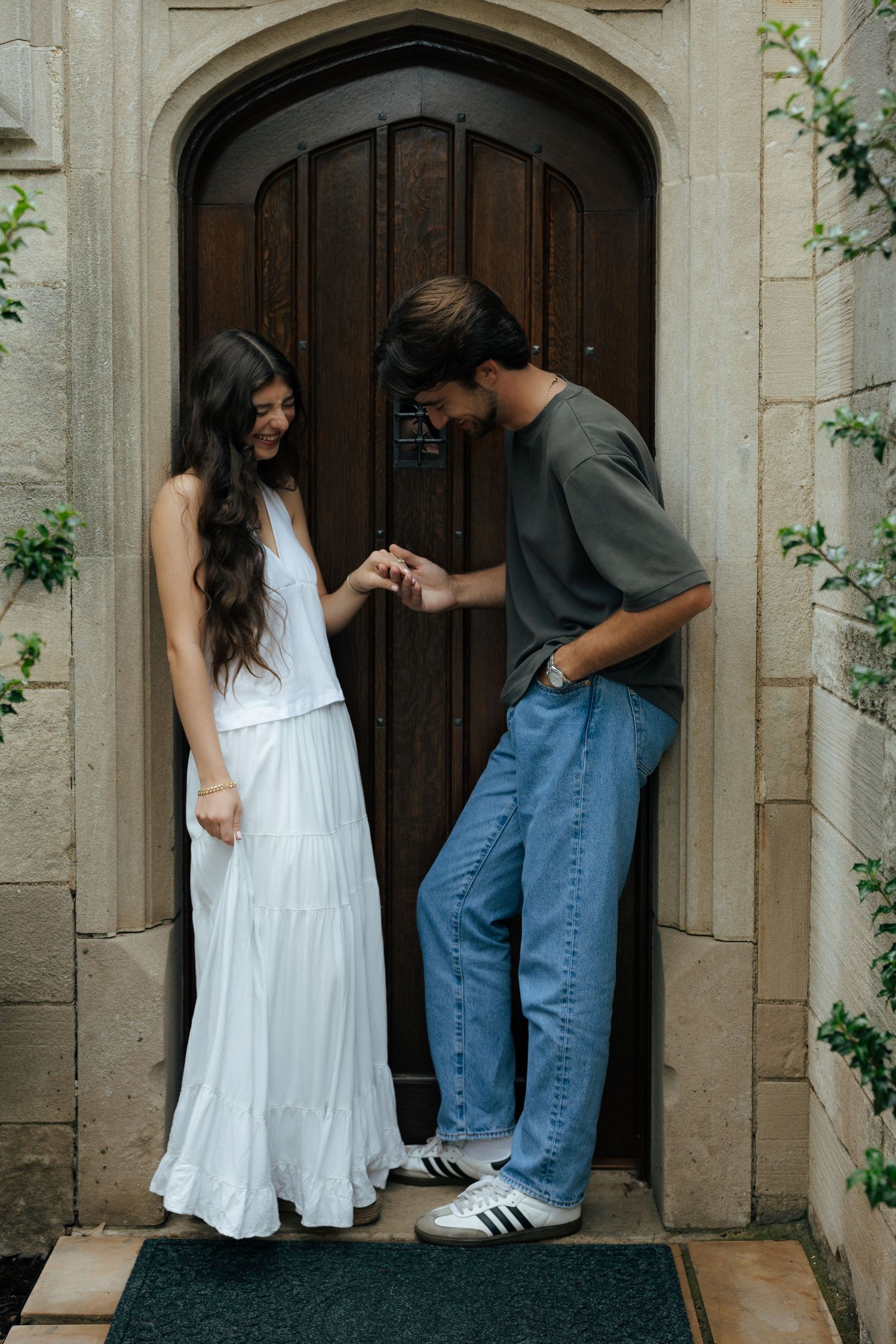 Couple sharing a romantic moment at an arched wooden door, woman in white dress holding hands with man.