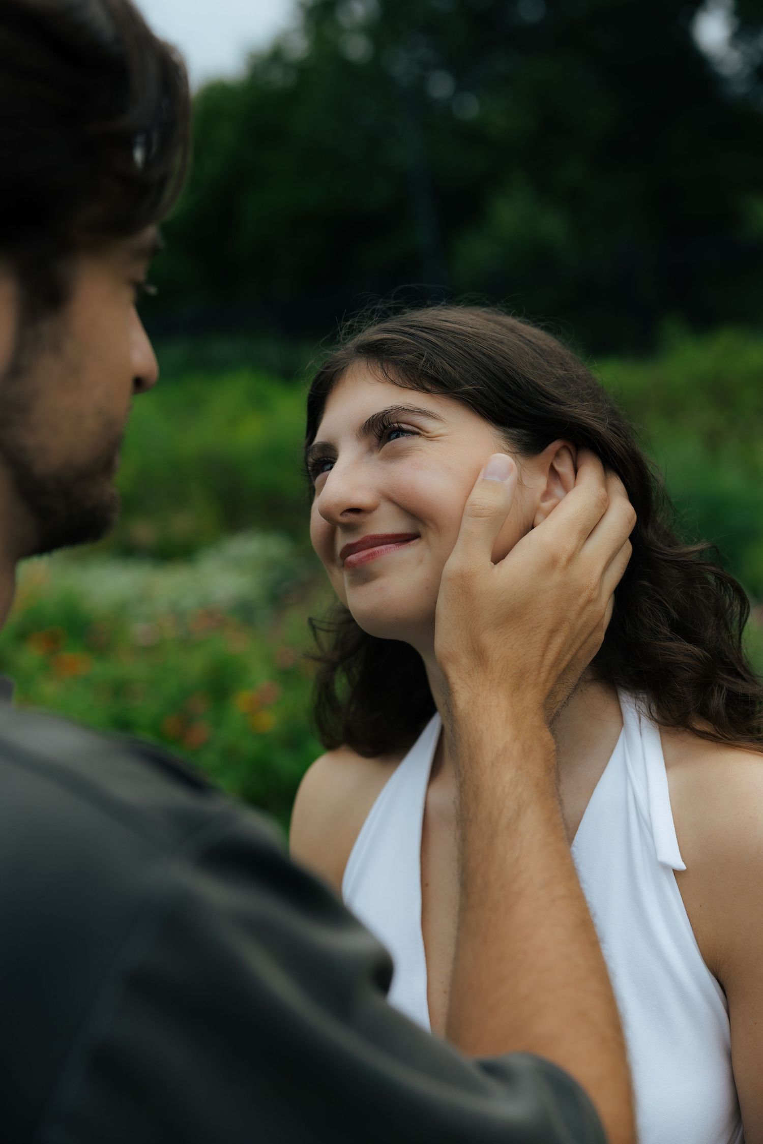 A man tenderly cups a smiling woman's face in a lush garden setting, sharing an intimate romantic moment outdoors.