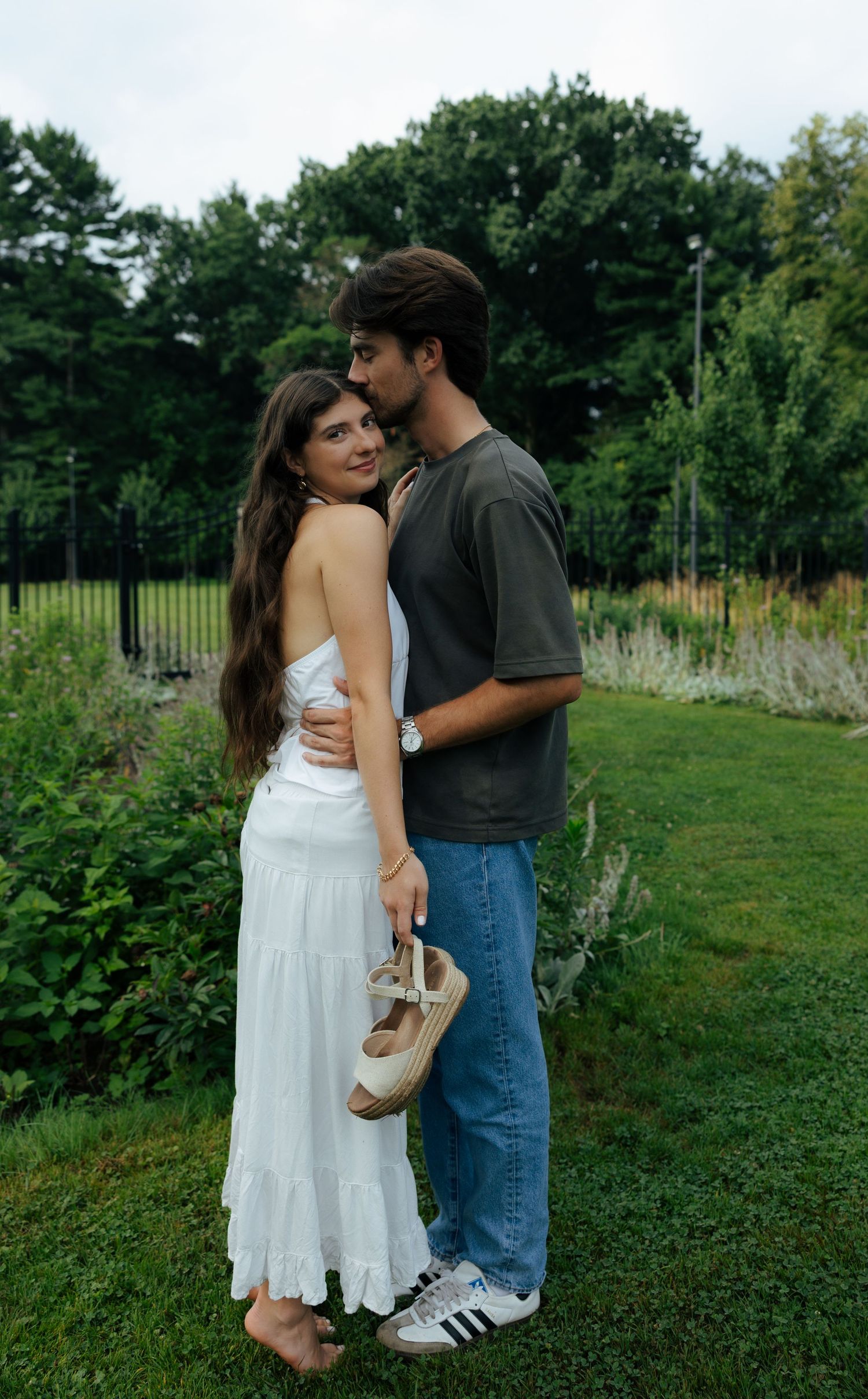 Couple embracing in a lush green garden, woman in white dress holding espadrille shoes barefoot.