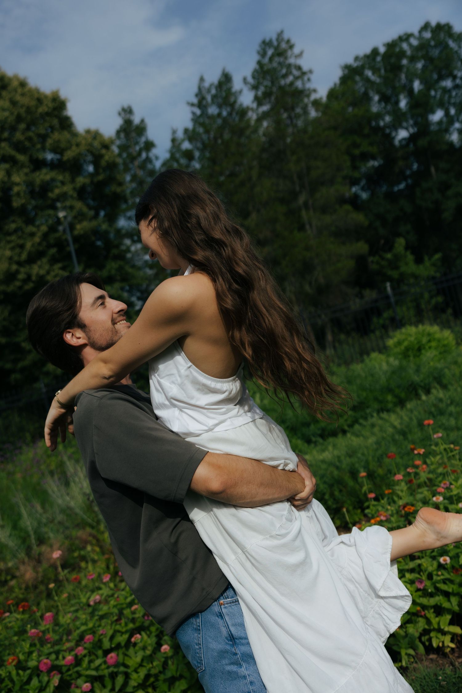 Man lifting woman in white dress joyfully in lush green garden during outdoor engagement session.