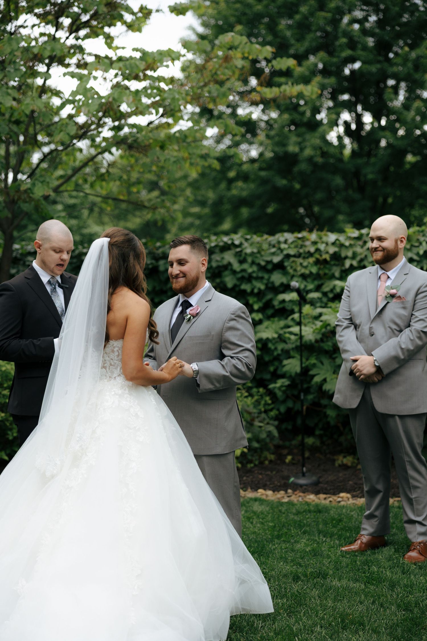 Groom smiles at bride during outdoor wedding ceremony with officiant and groomsmen.