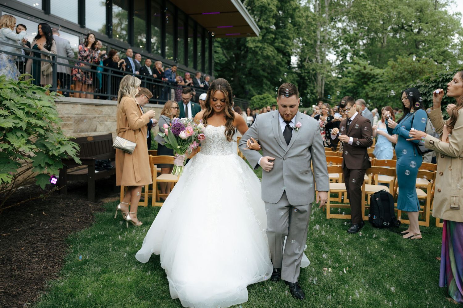 Bride and groom walk down aisle after outdoor wedding ceremony as guests celebrate with bubbles.