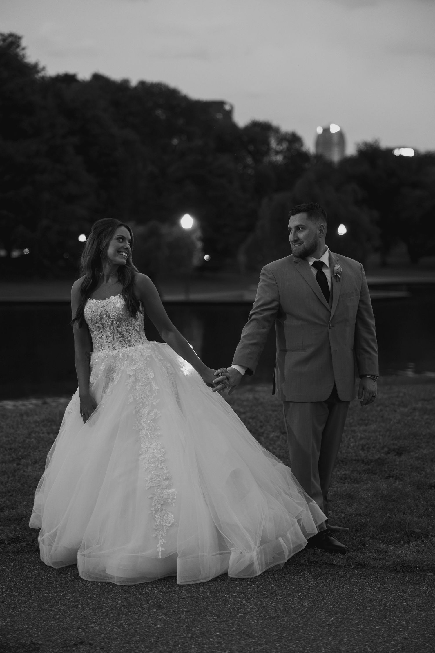 Black and white photo of smiling bride and groom holding hands walking outdoors at dusk.