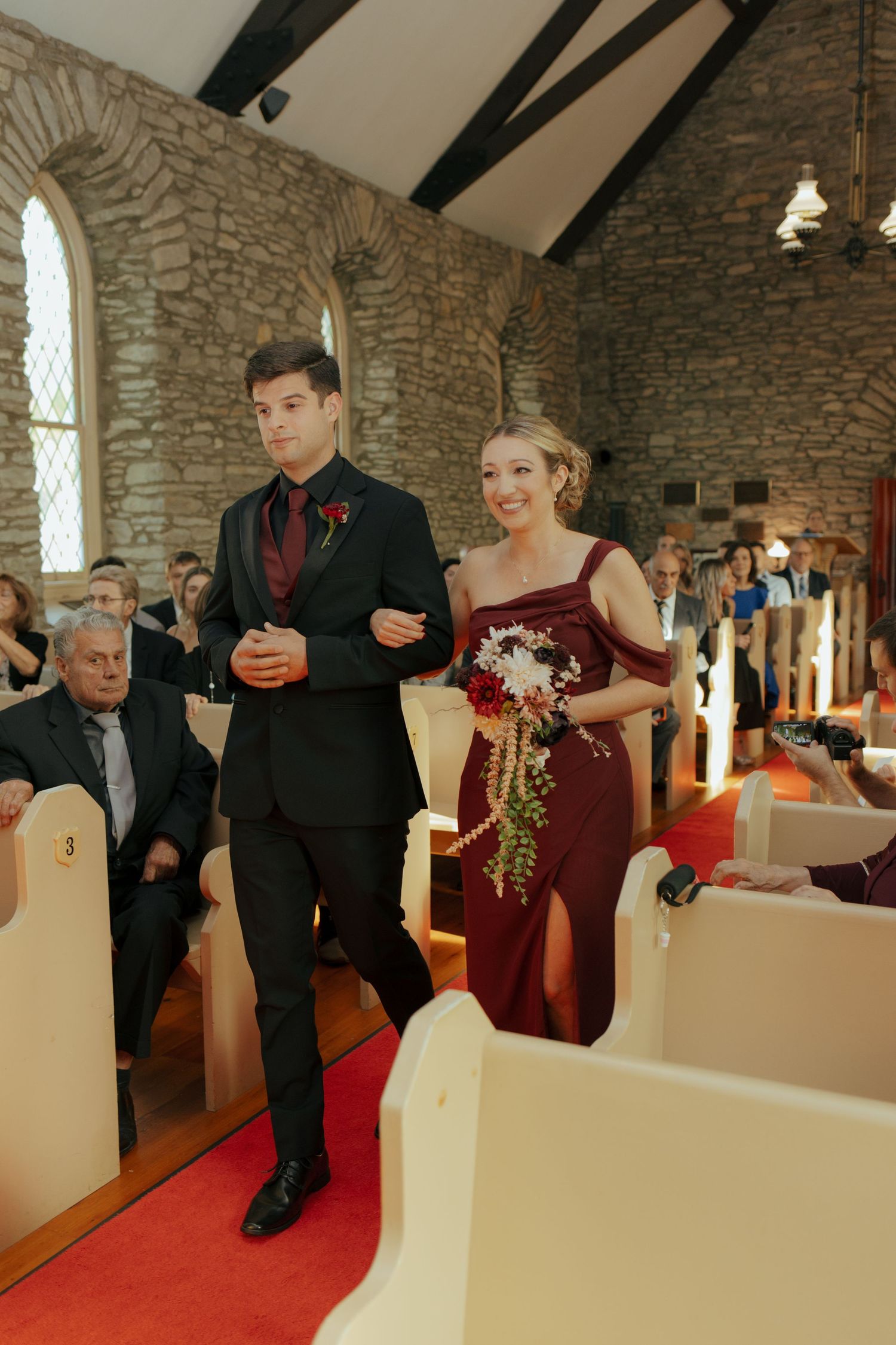 Groomsman in black tuxedo escorts bridesmaid in burgundy gown down red carpet aisle of stone chapel.