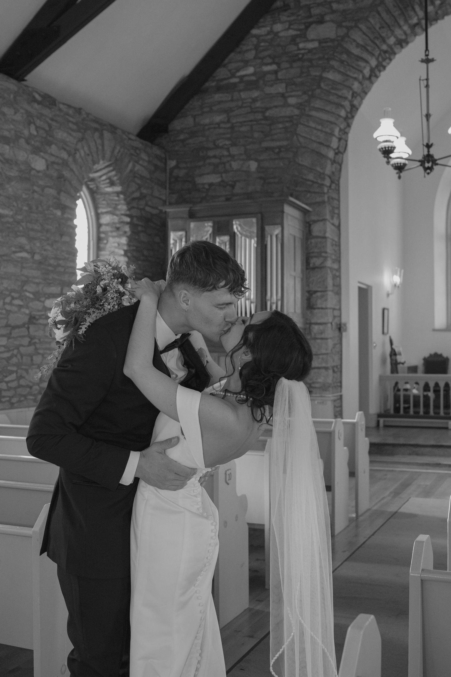Black and white photo of newlyweds sharing passionate kiss inside stone arch church with wooden pews in background.