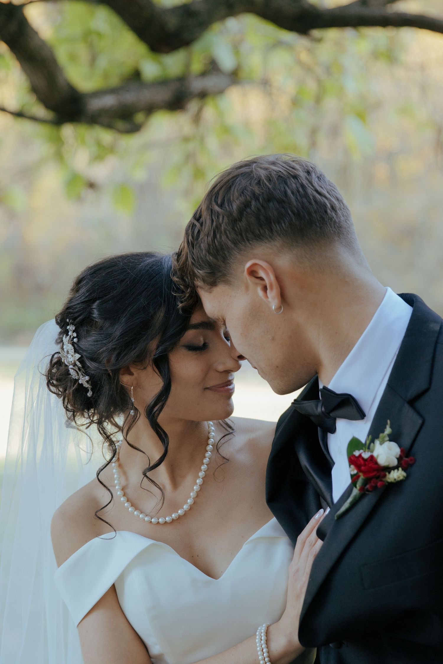 Bride and groom touching foreheads tenderly outdoors under autumn trees in an intimate wedding portrait.