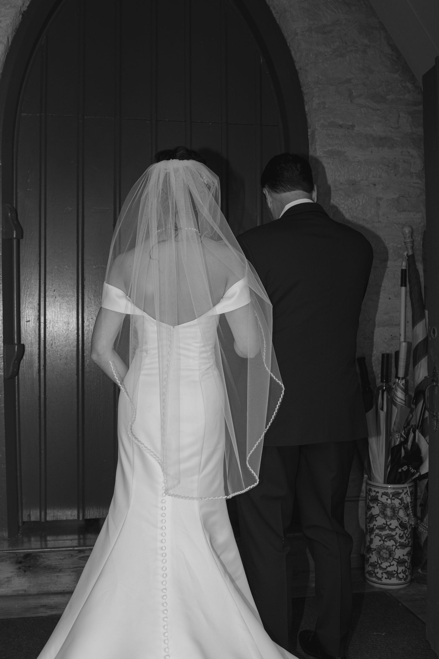 Bride in fitted white gown with veil and groom in black suit walk toward church door in black and white photo.