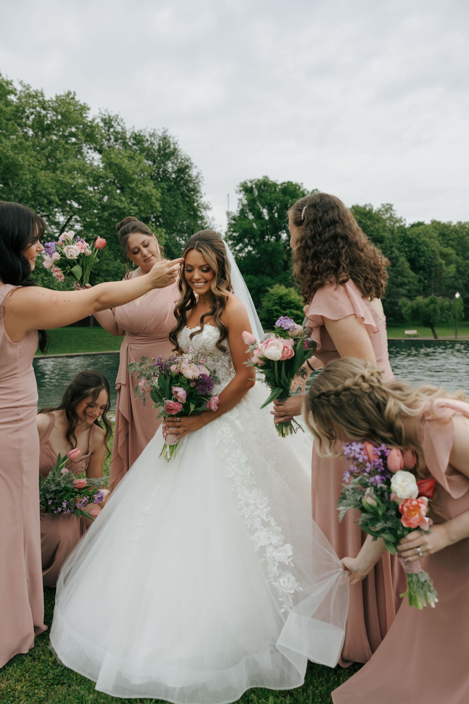Bride in white gown surrounded by bridesmaids in blush pink dresses holding floral bouquets outdoors near a pond.