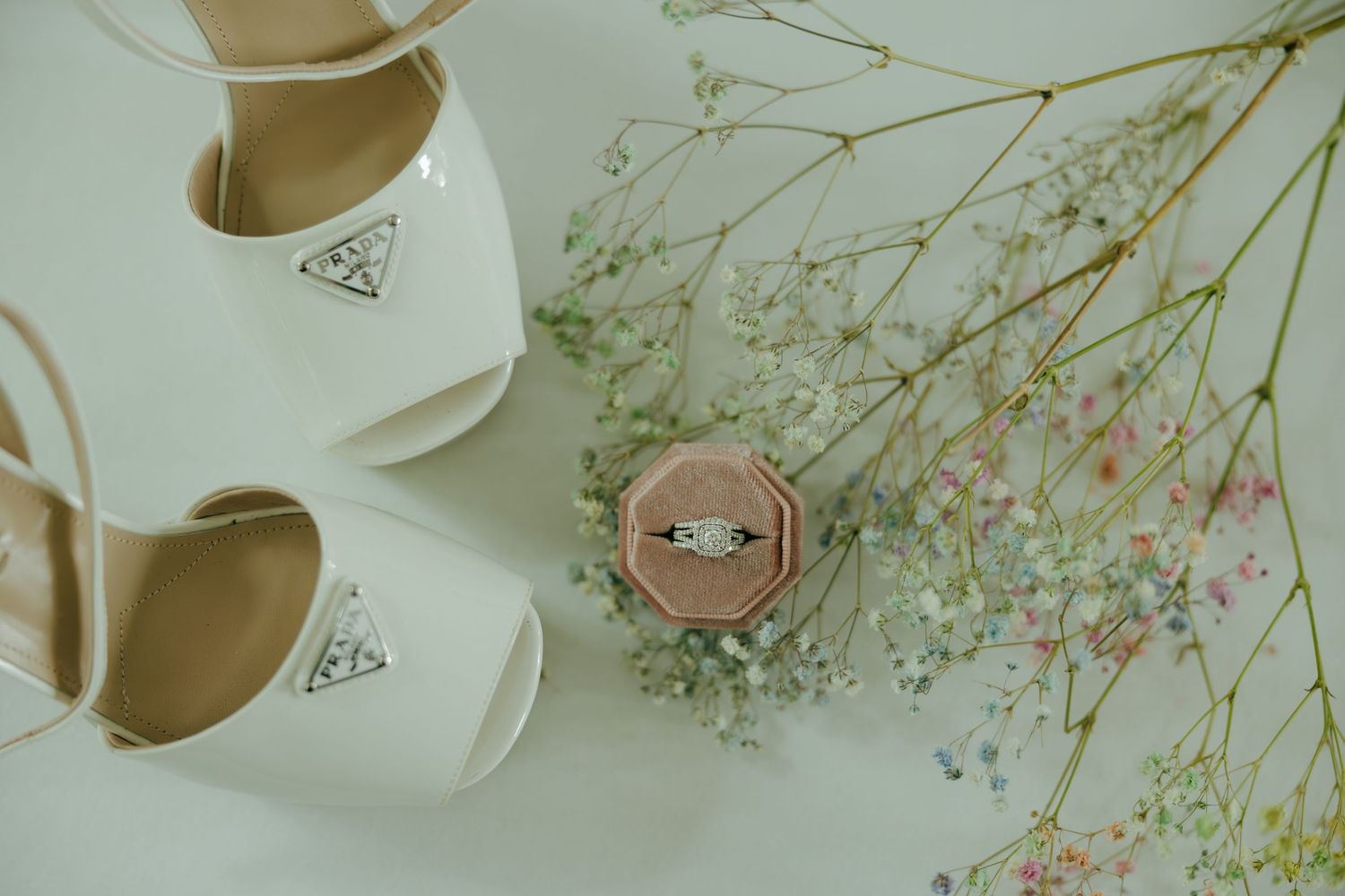 White bridal heels and diamond engagement ring in velvet box with delicate baby's breath flowers on white background.