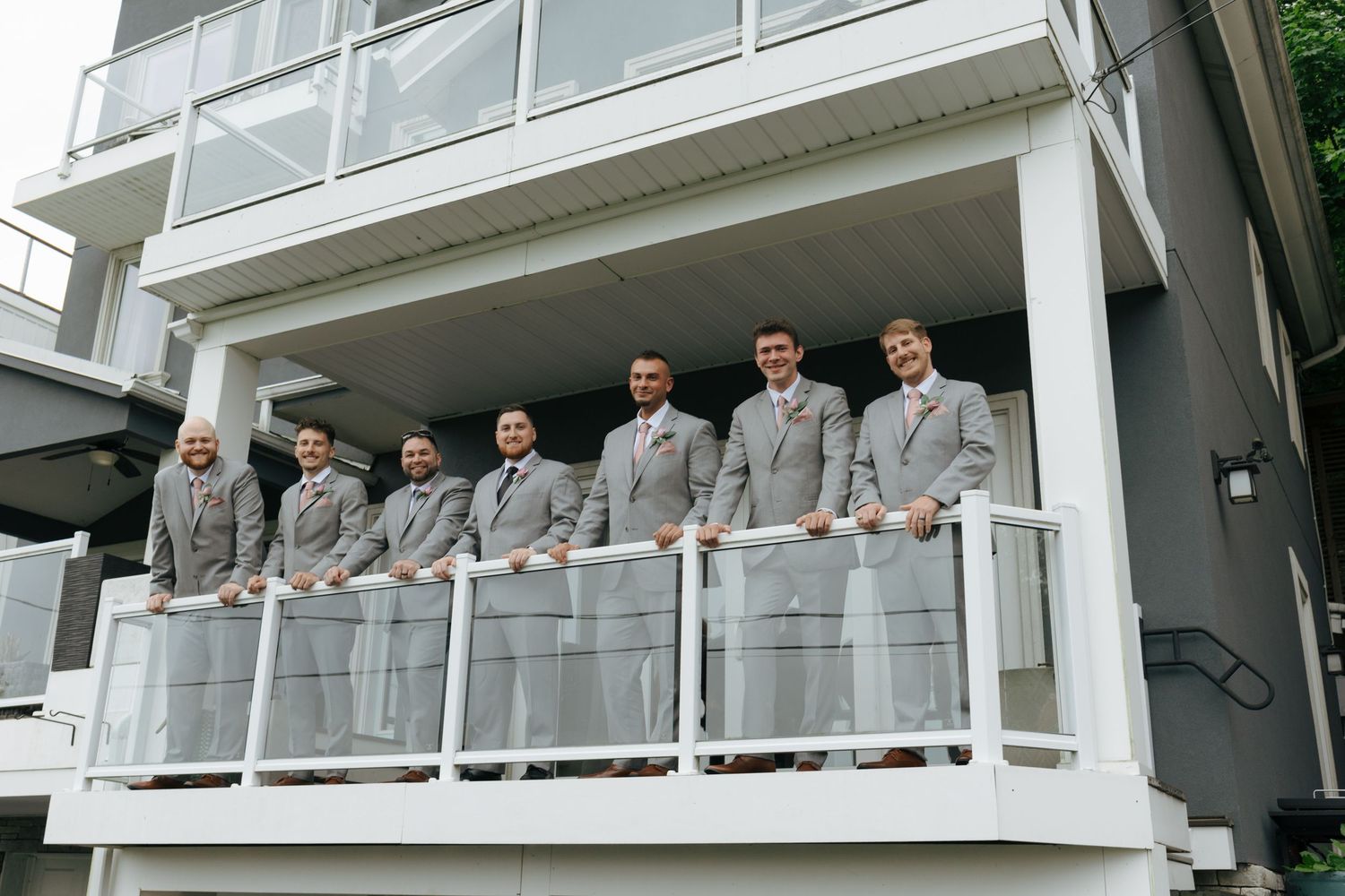 Groomsmen in matching gray suits pose together on a white balcony railing outside a modern building.