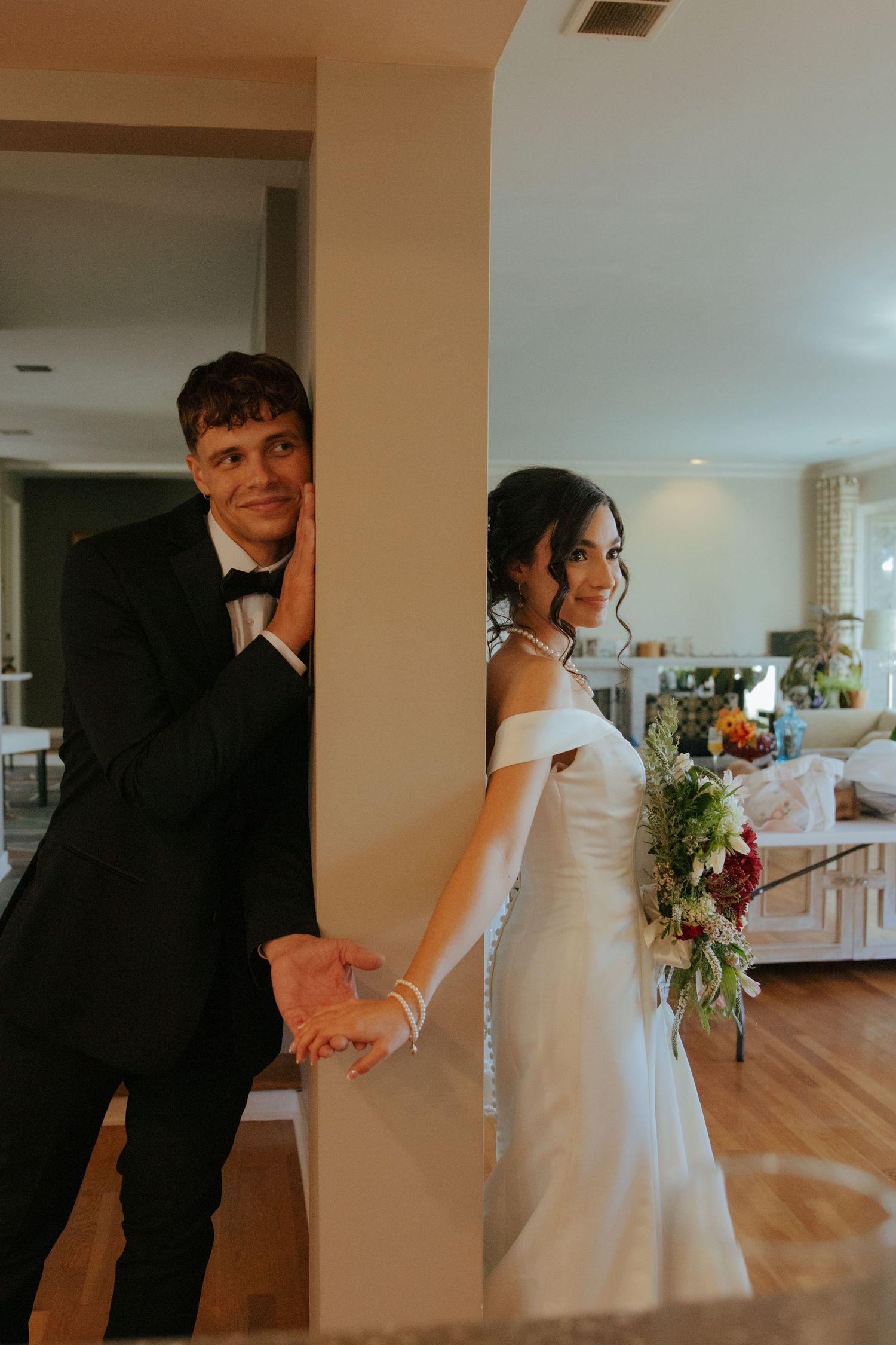 Bride and groom holding hands around a pillar, smiling, bride holding colorful bouquet.