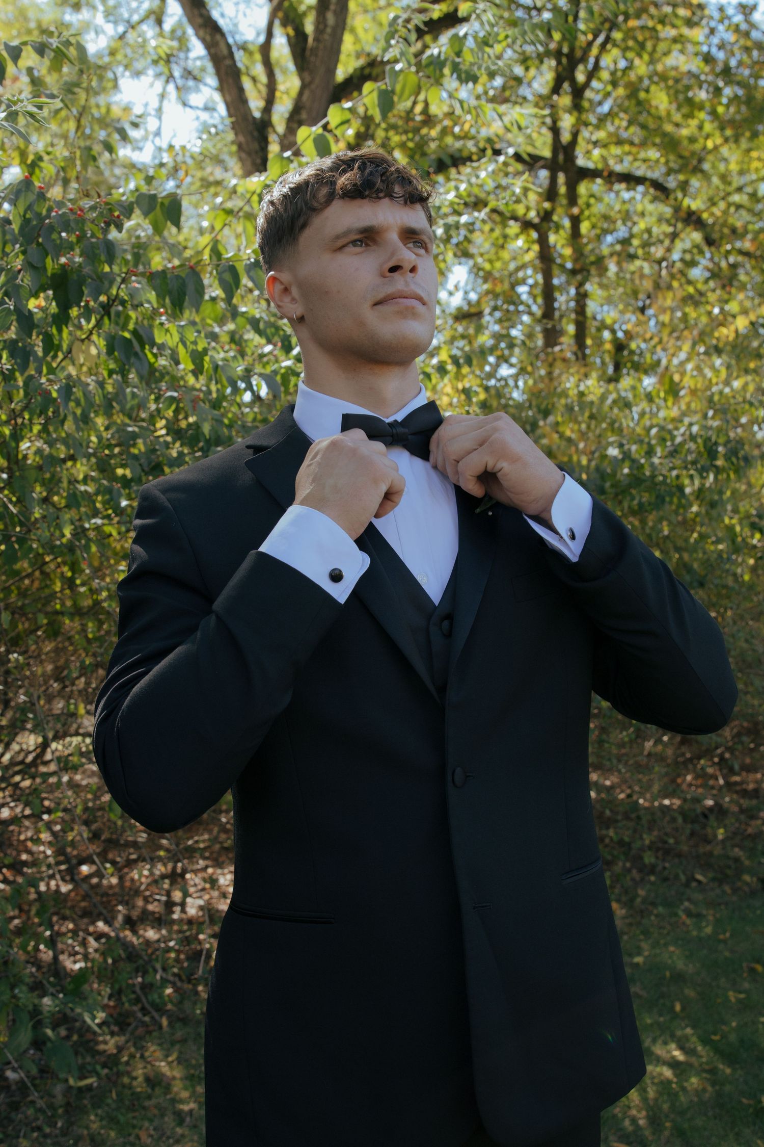 Young man in black tuxedo adjusting bow tie outdoors among green trees.