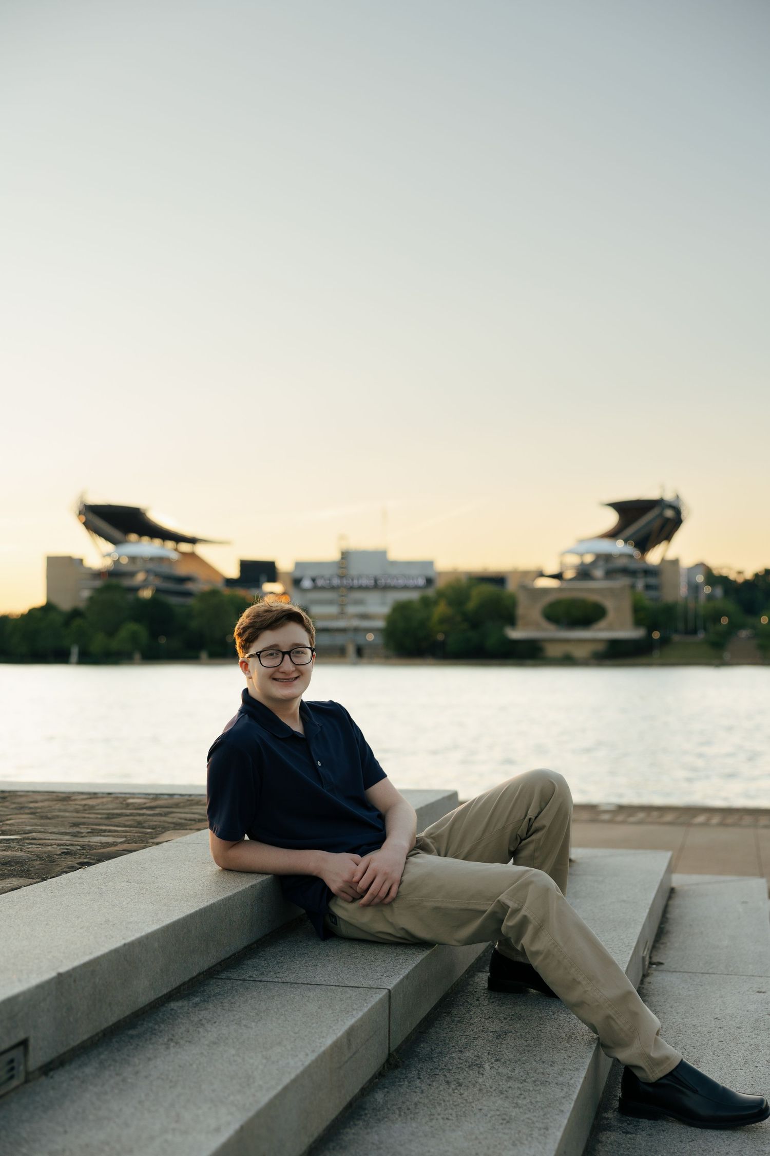 Teen boy in navy polo sitting on riverside steps with Acrisure Stadium visible across the water.