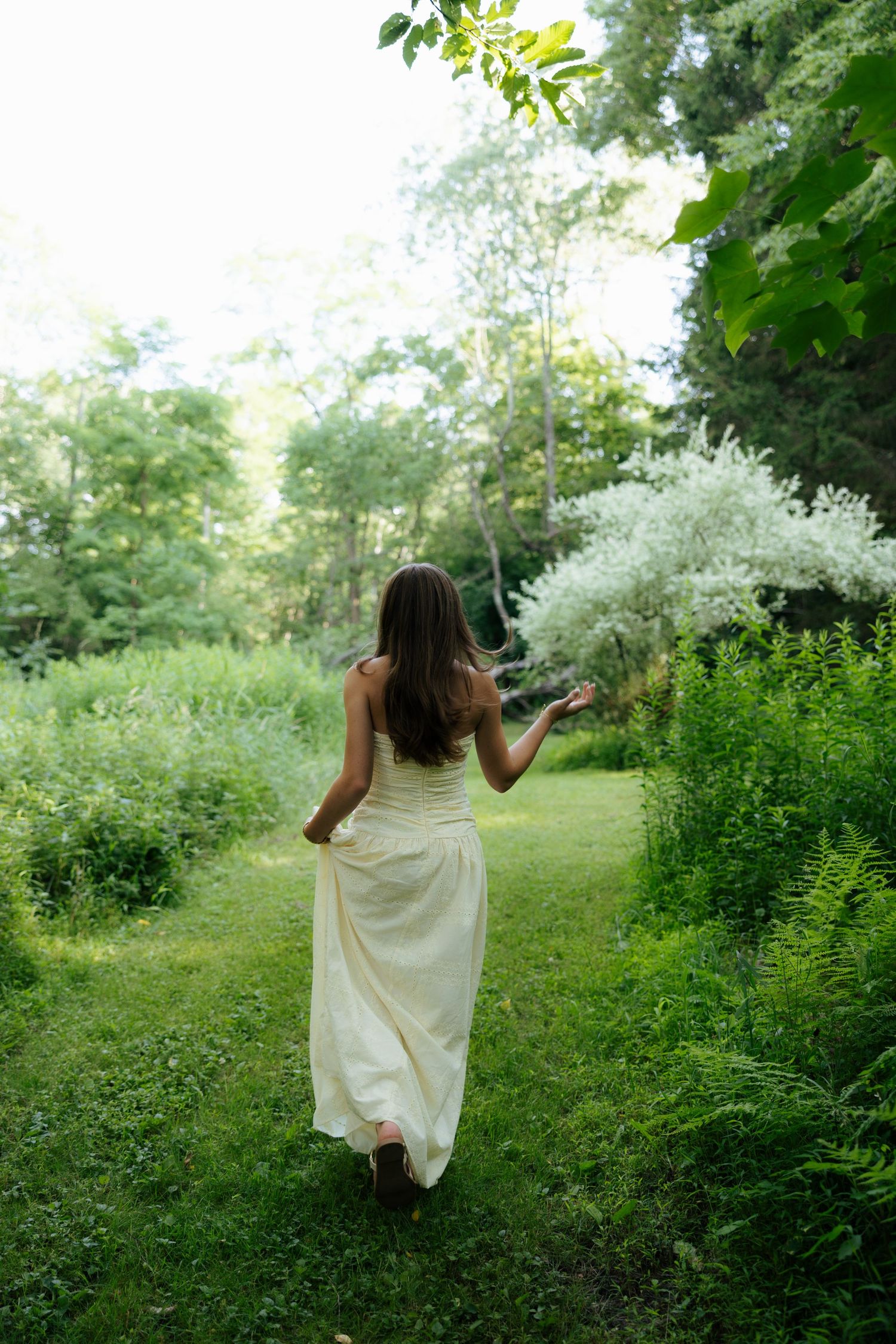 Woman in flowing white dress walking through a lush green garden path toward blooming white flowers.