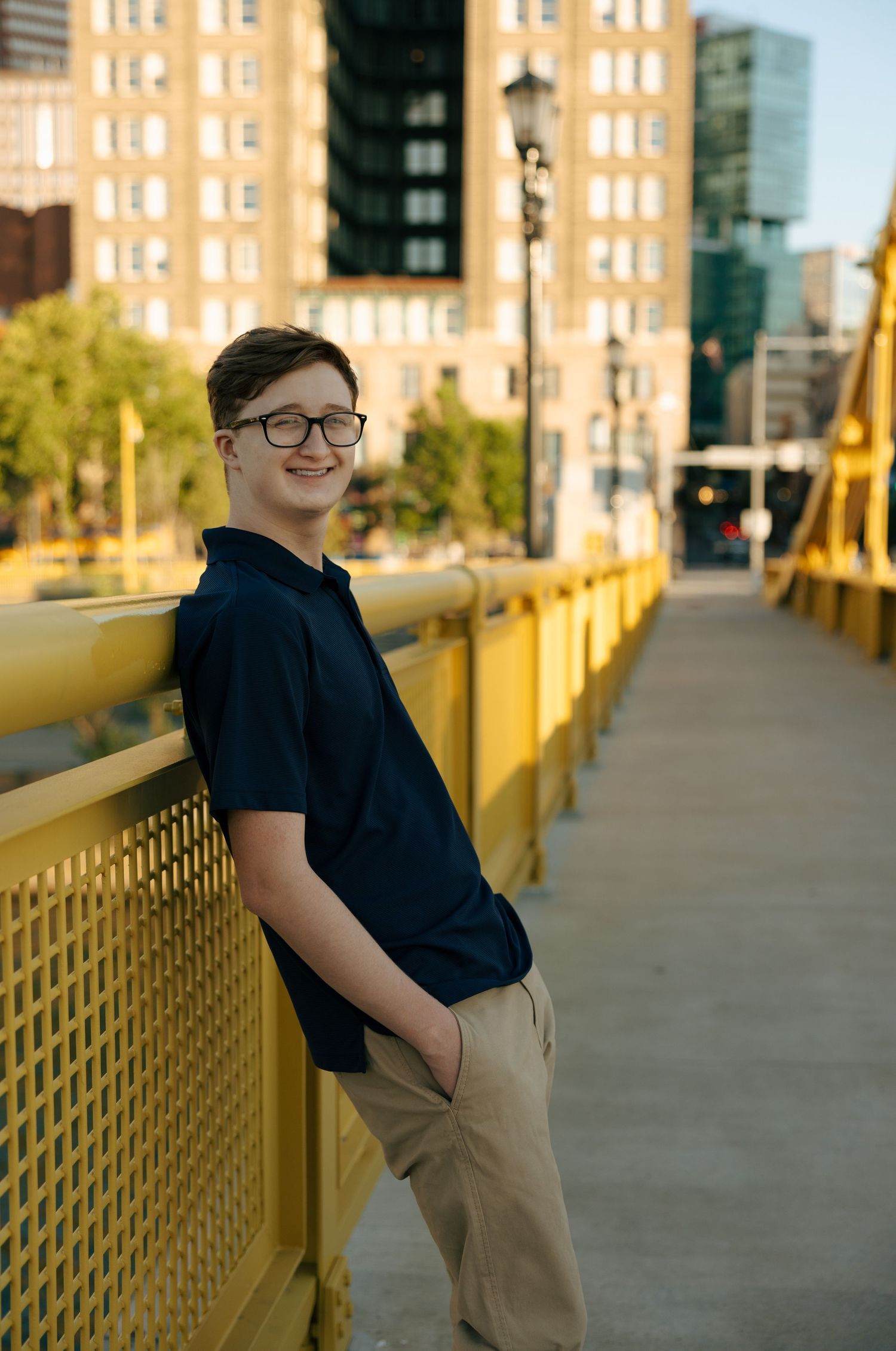 Teen boy in navy polo and khakis leaning on yellow bridge railing with Pittsburgh city buildings in background.