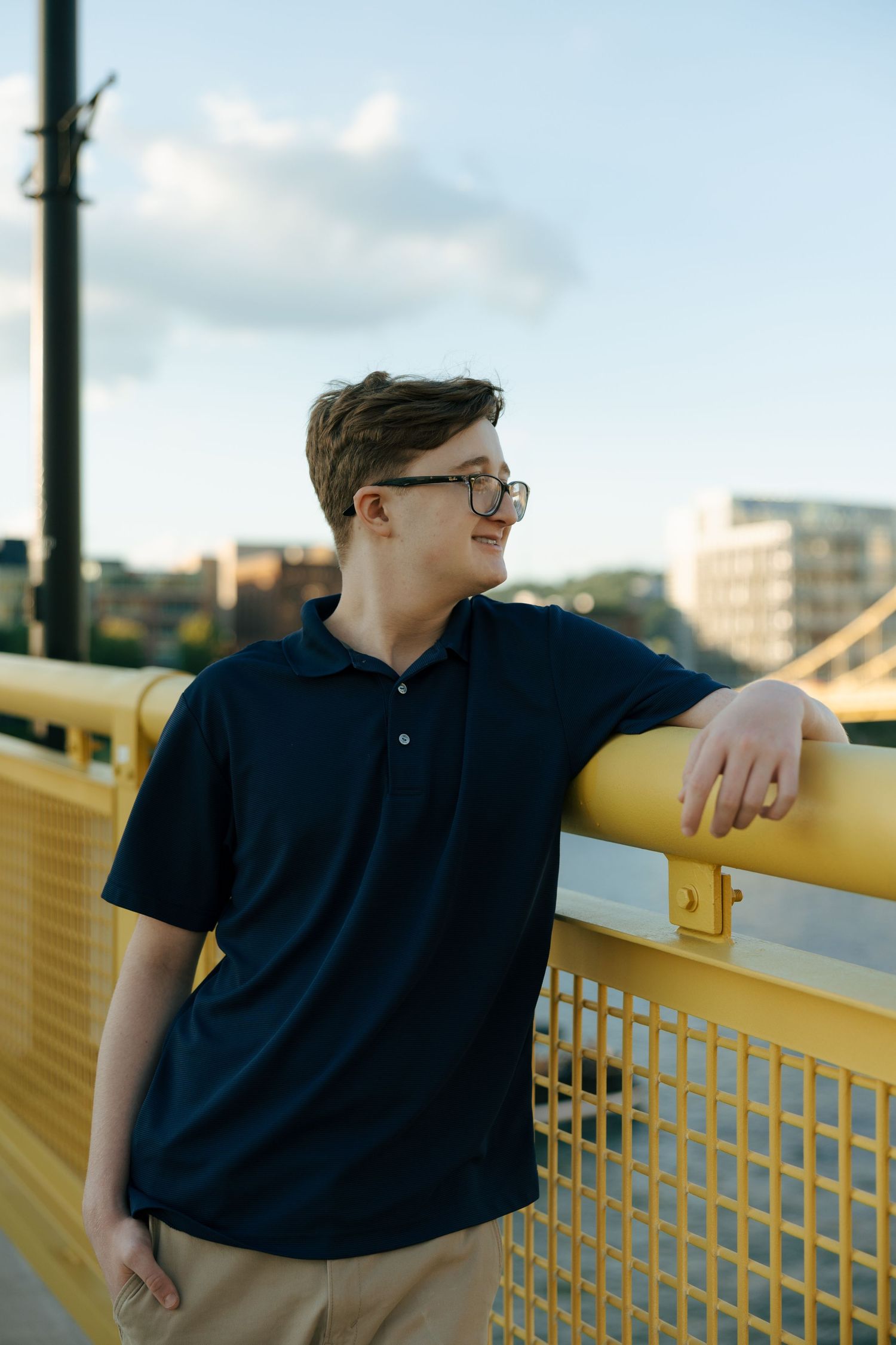 Smiling teen in navy polo shirt leaning on yellow bridge railing with urban skyline backdrop.