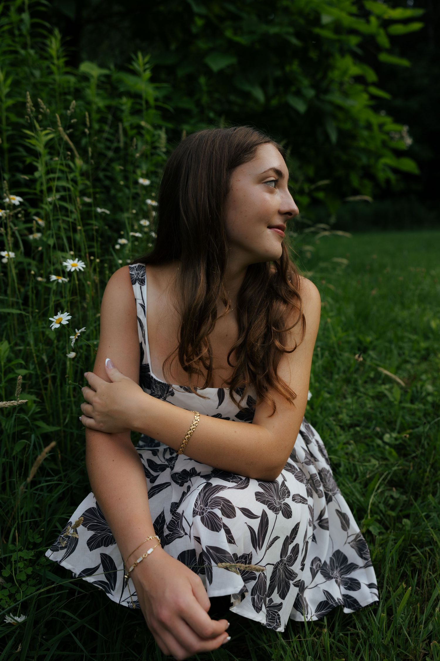 Young woman in floral dress sitting in green meadow with daisies, smiling outdoors.