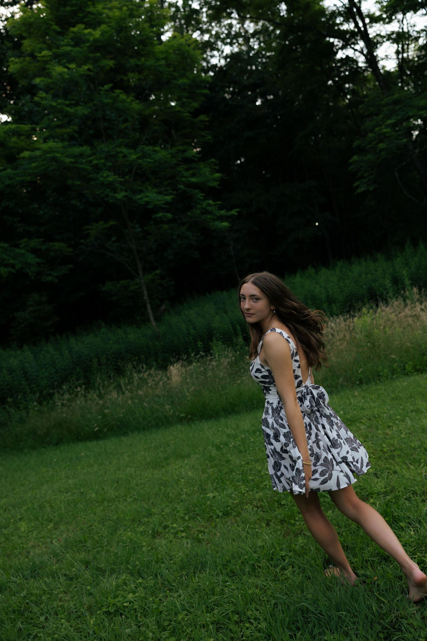 Young woman in floral dress twirling barefoot on lush green grass with trees in background.