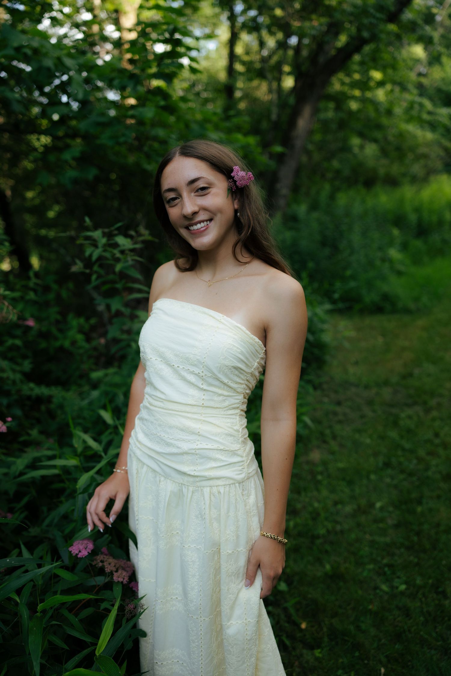 A joyful young woman in a white strapless dress stands among pink flowers in a lush garden.