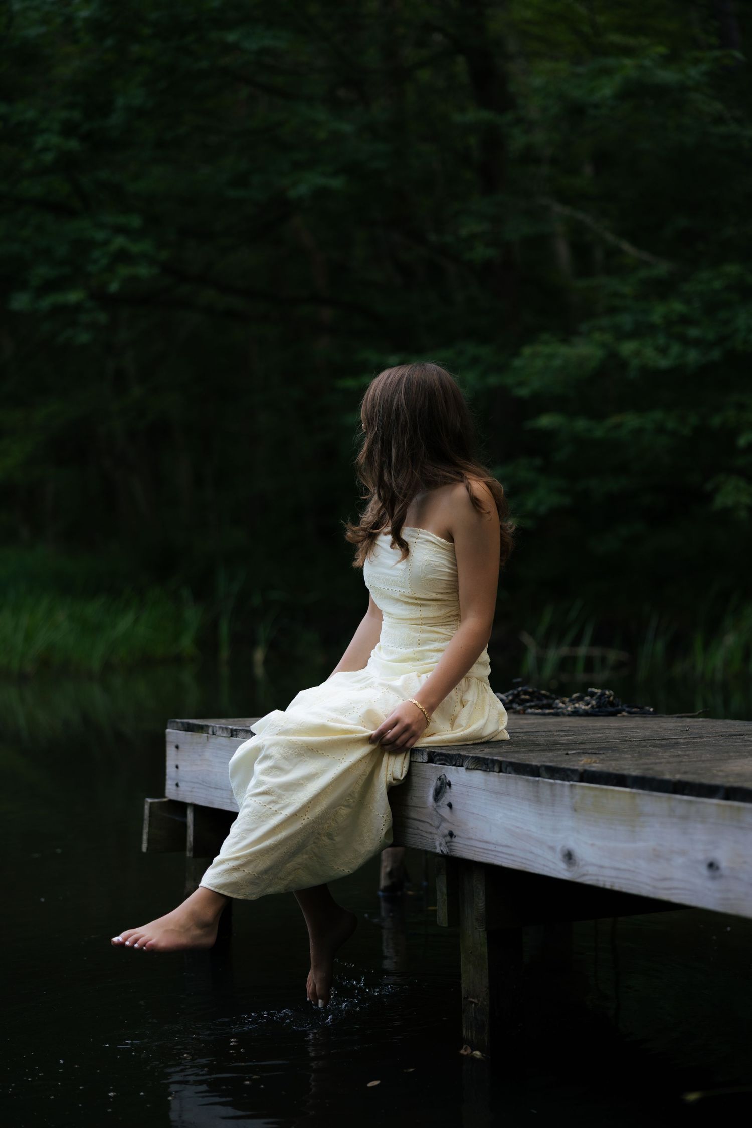 Young barefoot woman in white dress sitting on wooden dock over dark lake at dusk.