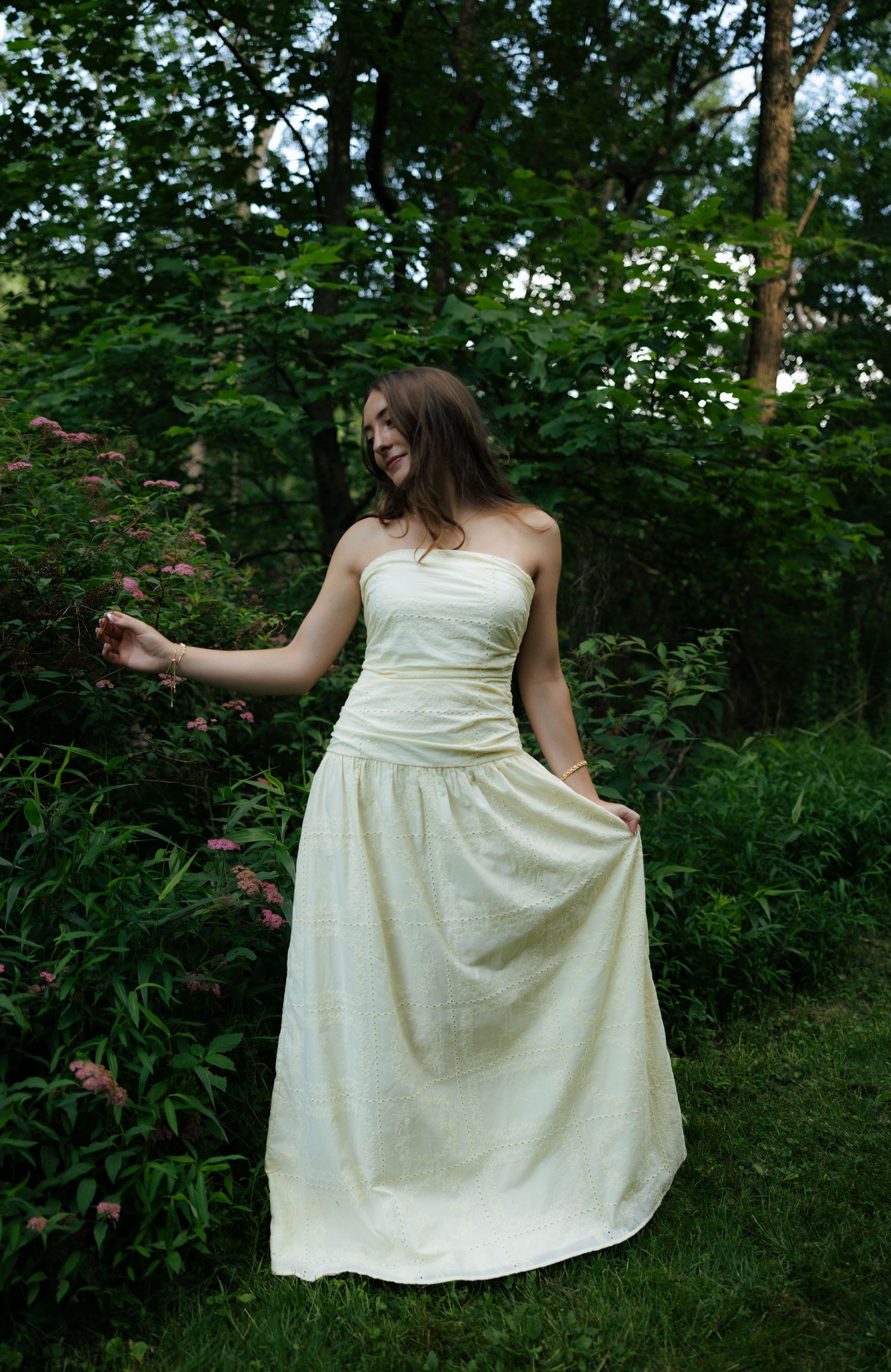 Young woman in strapless cream ball gown standing among green garden foliage, gently touching pink flowers.