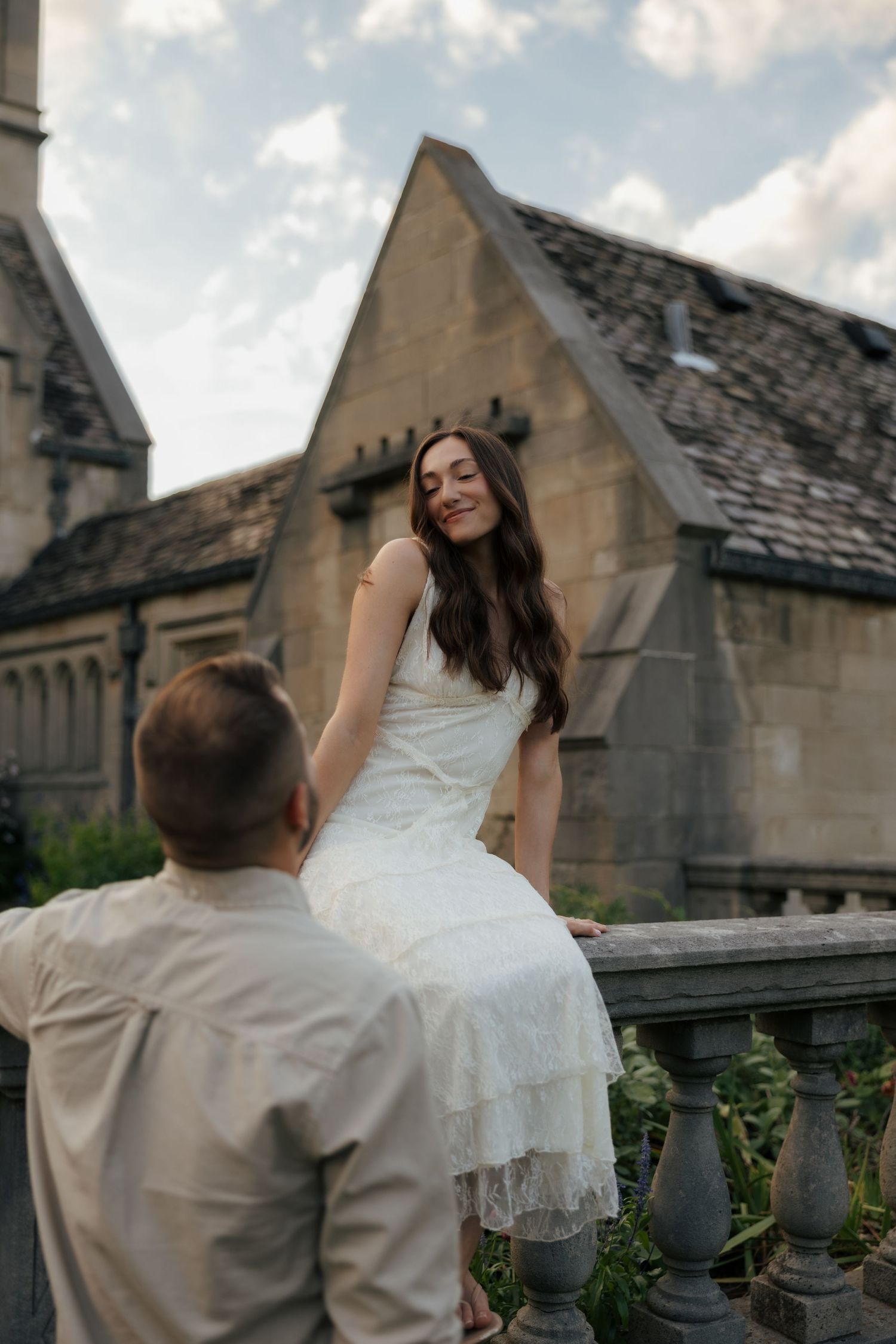Bride in white lace dress sits on stone balustrade smiling at groom before historic building.