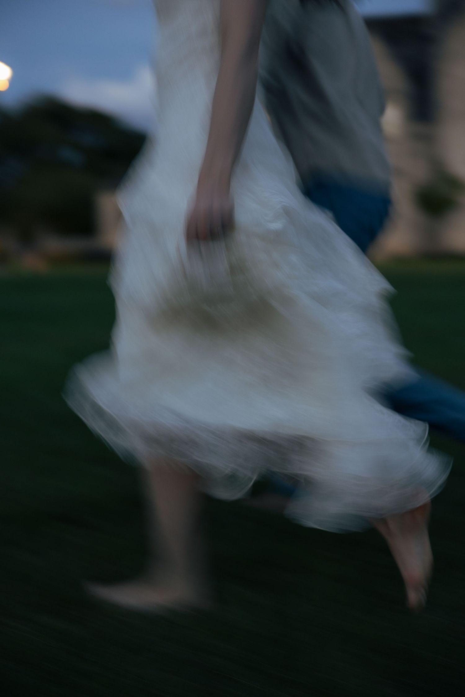 A motion-blurred image of a woman in a flowing white dress dancing outdoors at dusk.