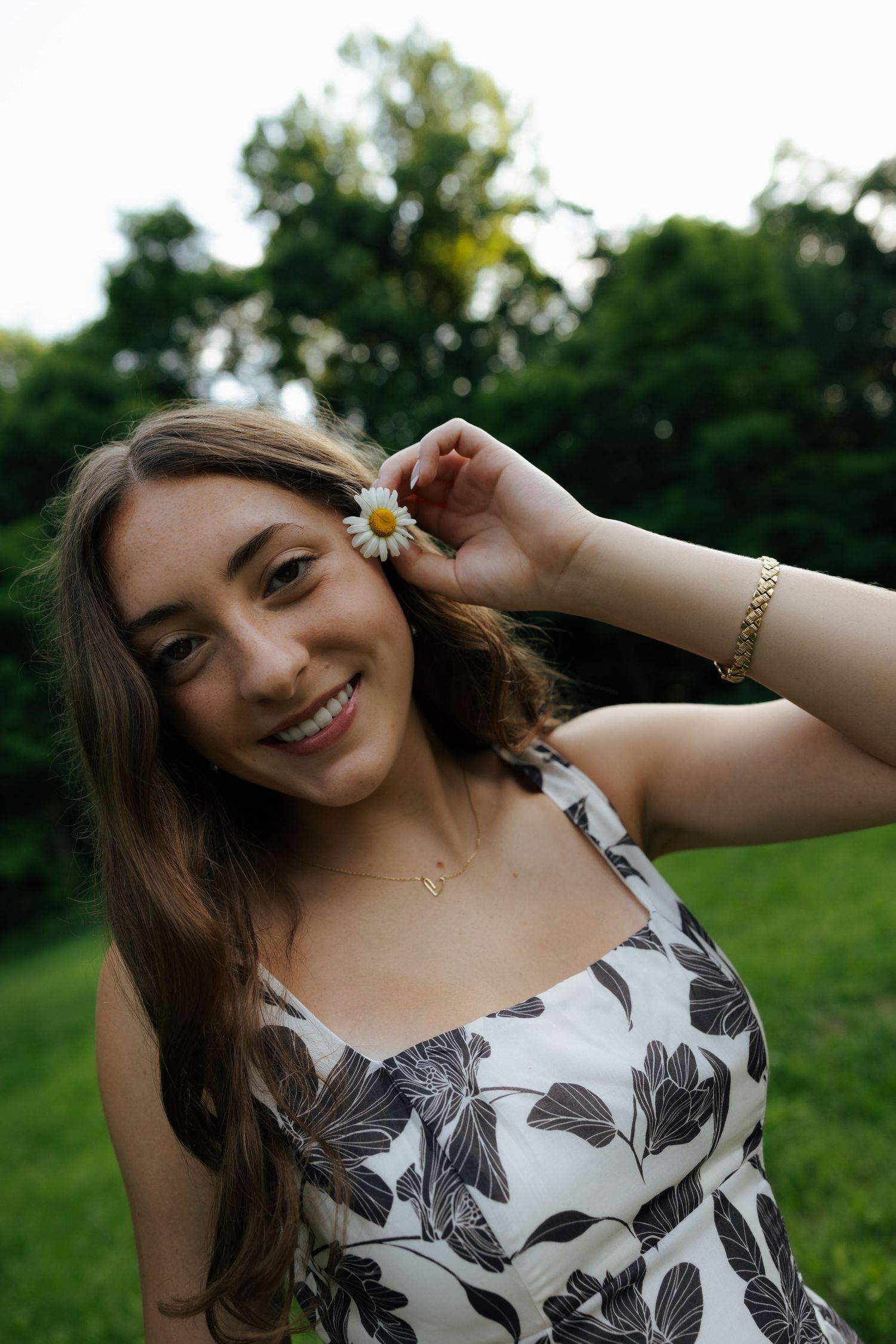 Smiling young woman in floral dress holding a daisy flower near her ear in a lush green park.